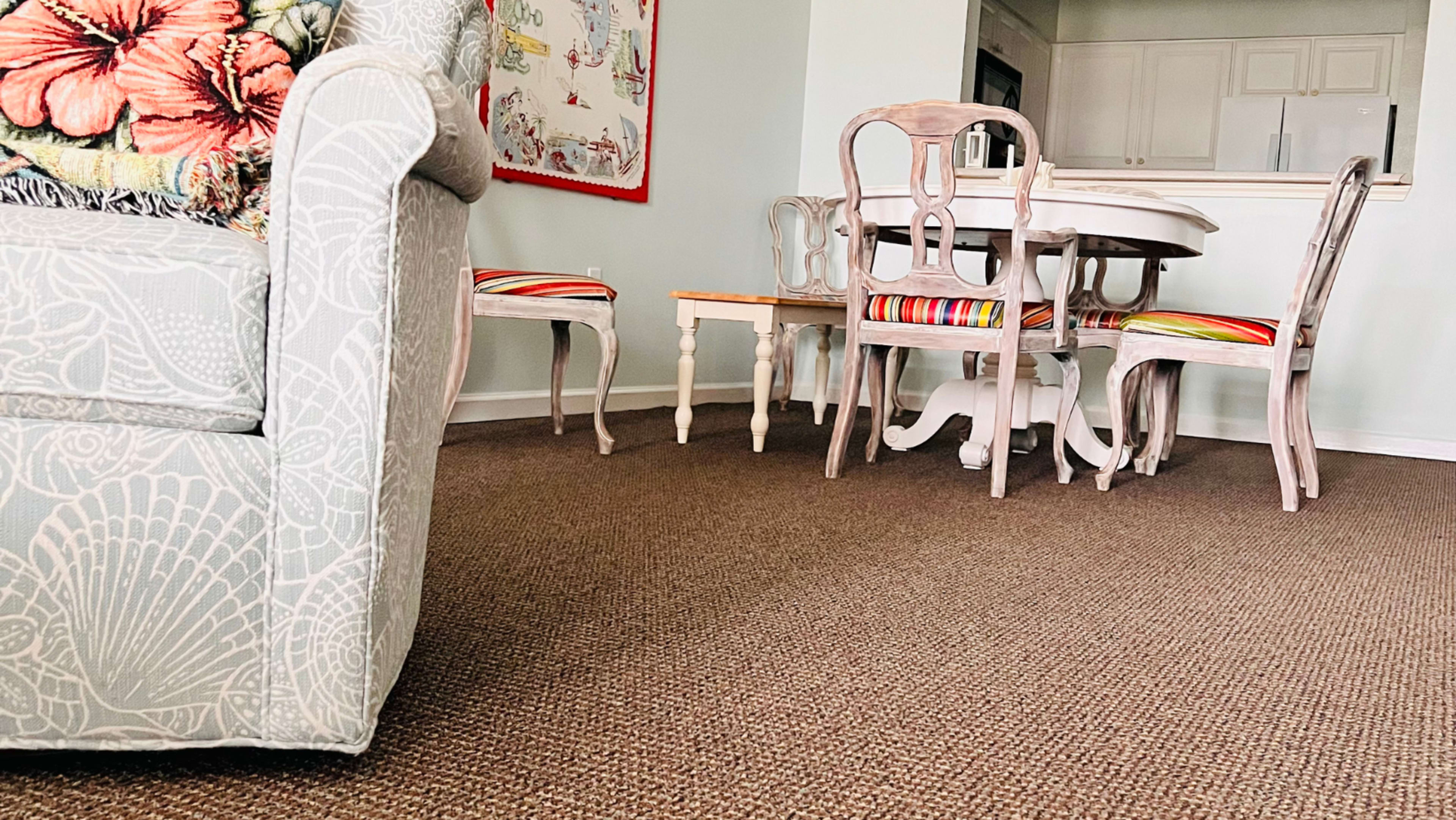A light-colored sofa with floral patterns is positioned near a wooden dining table surrounded by chairs with striped cushions in a room with beige carpet.