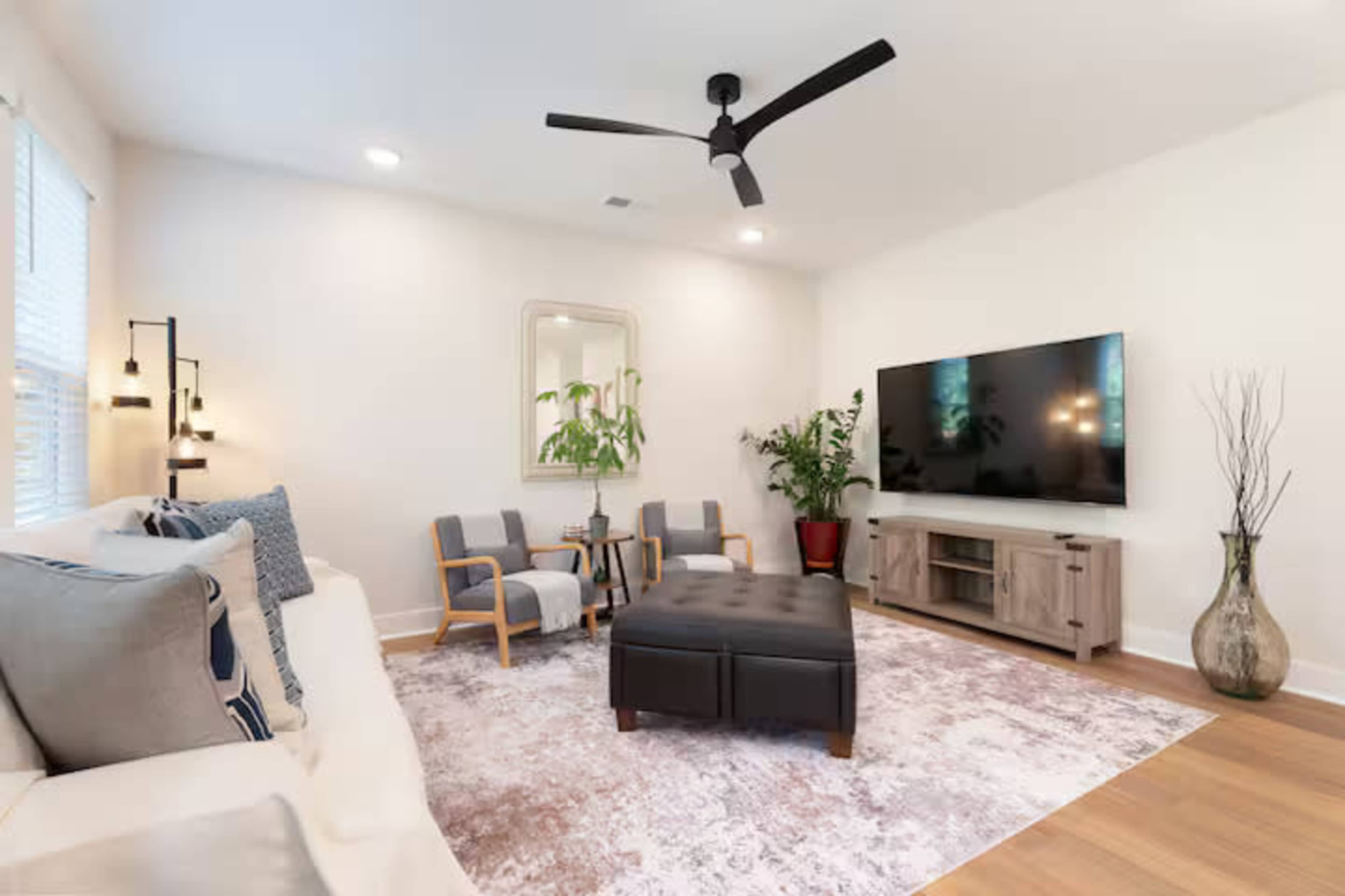 The living room features a light color scheme with a large TV on the wall, a dark ottoman, two armchairs, and a potted plant.