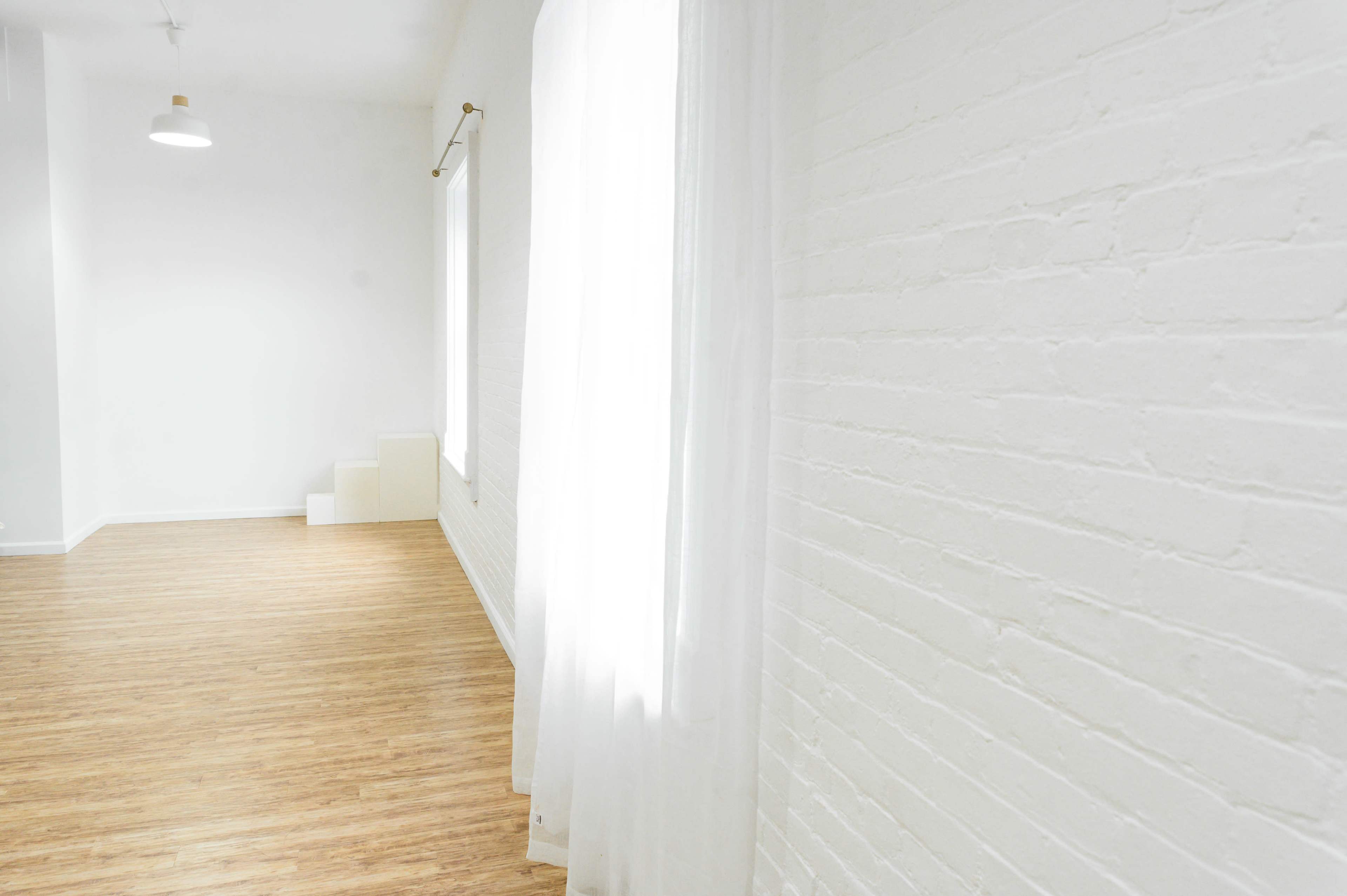 A bright, empty room with white brick walls, wooden flooring, and sheer curtains near a window.