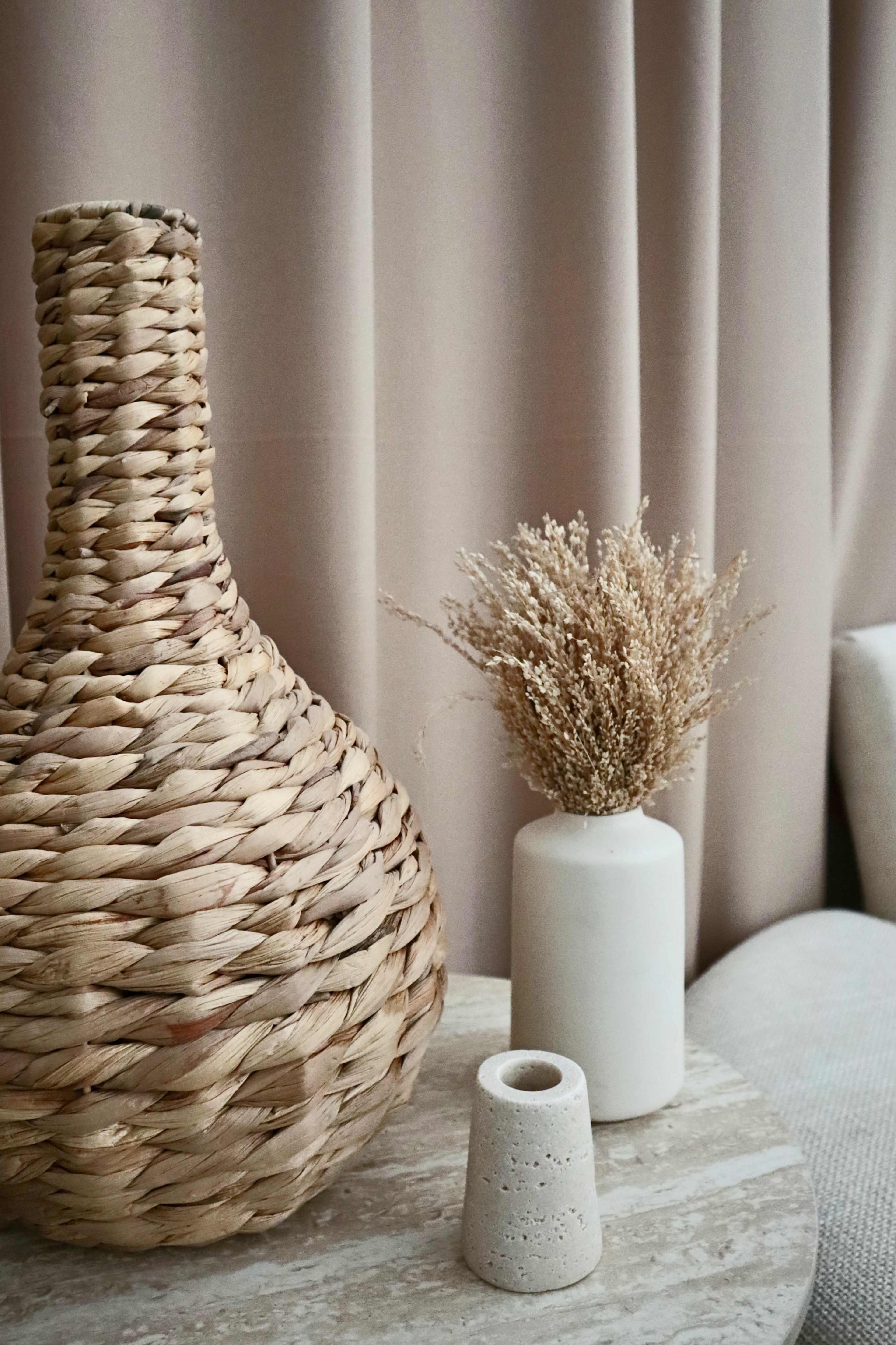 A woven basket-shaped vase sits on a table next to a white cylindrical vase filled with dried flowers and a small cylindrical concrete holder.