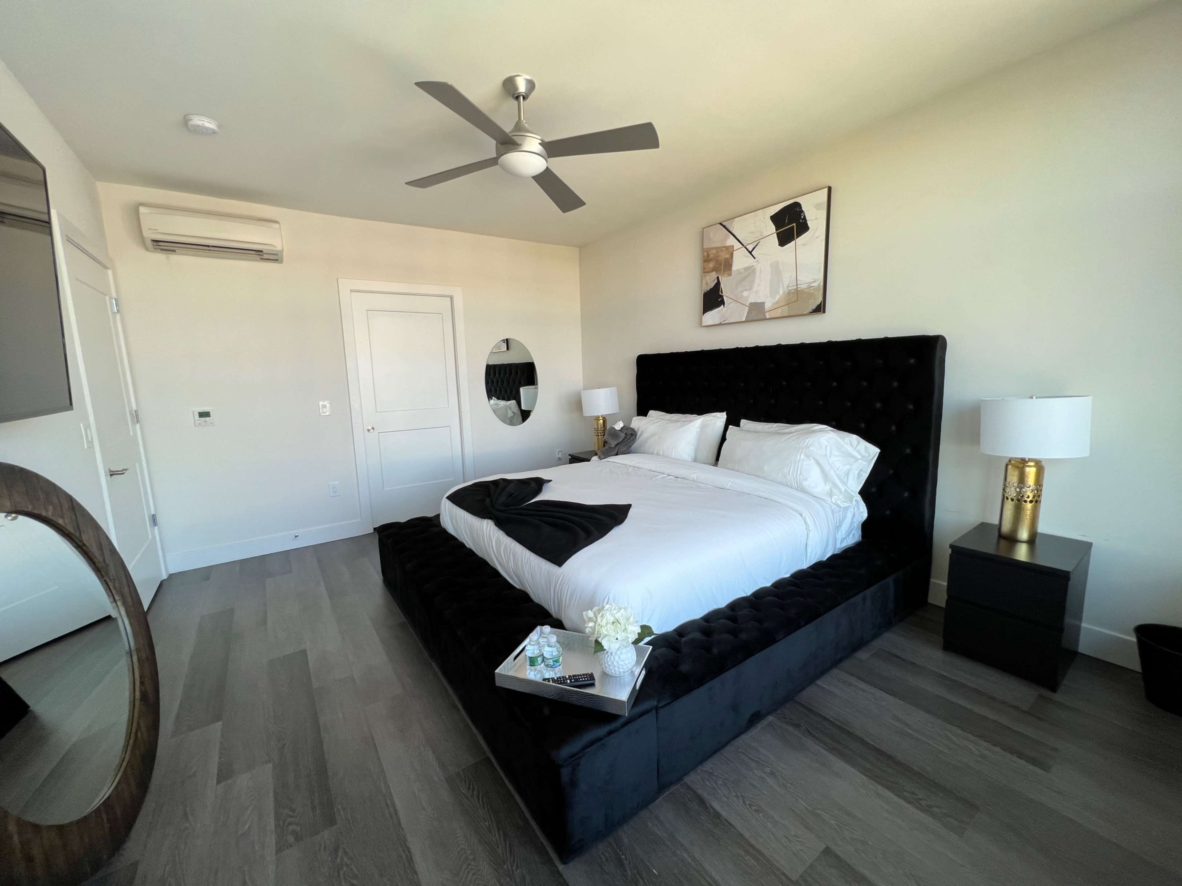 A modern bedroom featuring a large black upholstered bed, a round mirror, a nightstand with a lamp, and wooden flooring.
