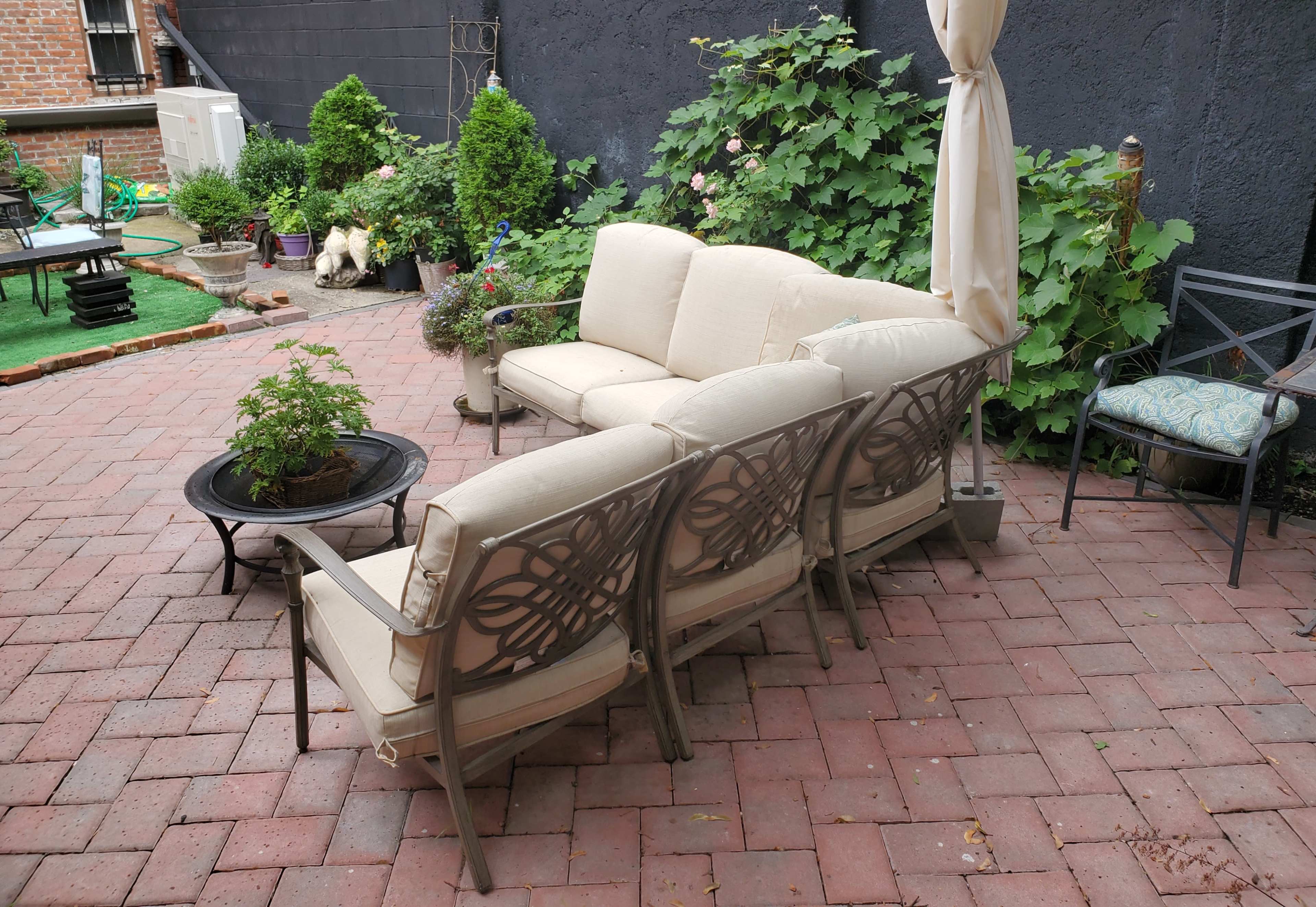 A patio area with a beige sofa set and armchairs arranged around a circular black firepit, surrounded by greenery and brick paving.