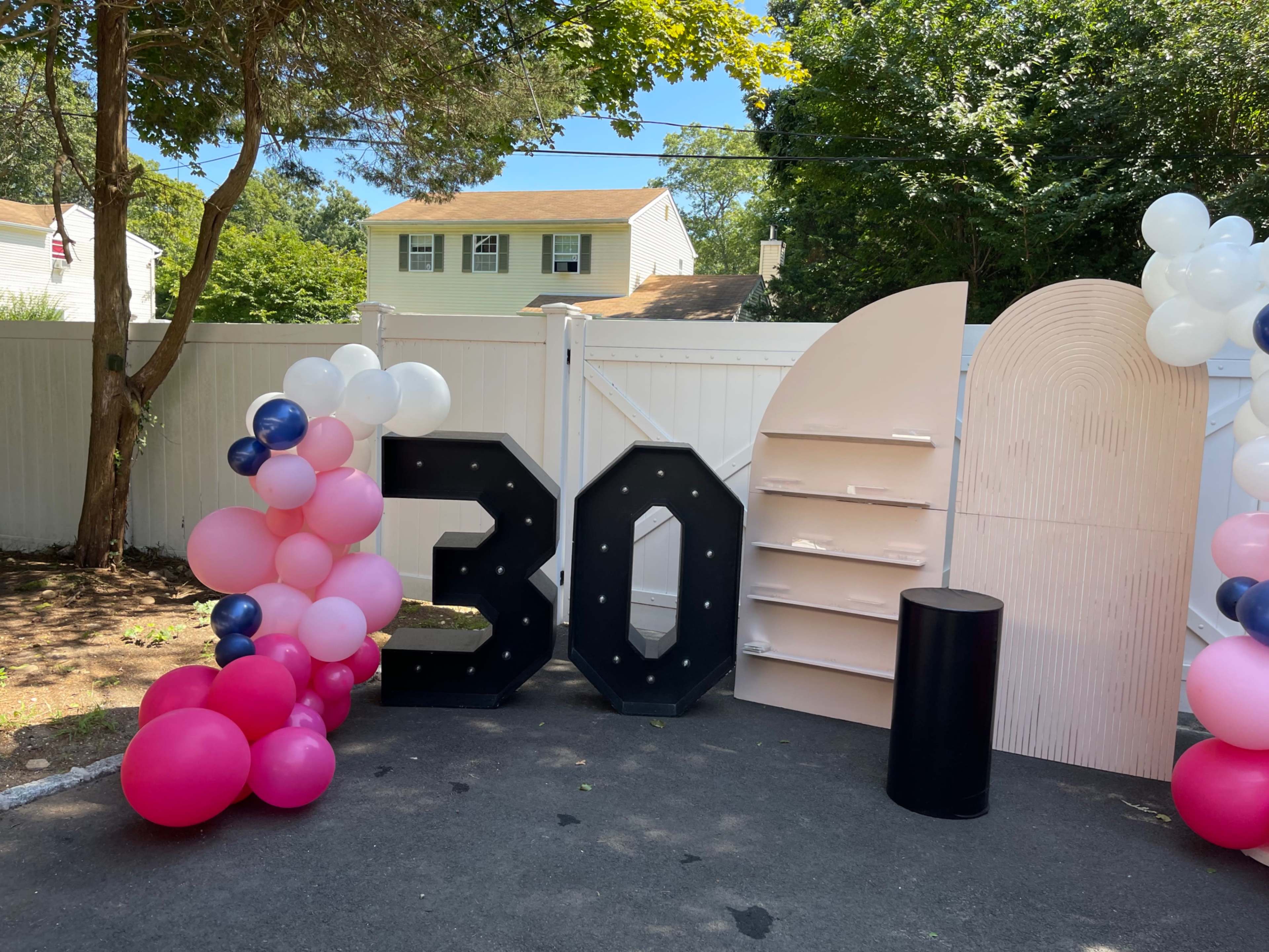 A decorated outdoor area features large black "30" letters flanked by pink and blue balloons, alongside a shelf unit and a black circular table.