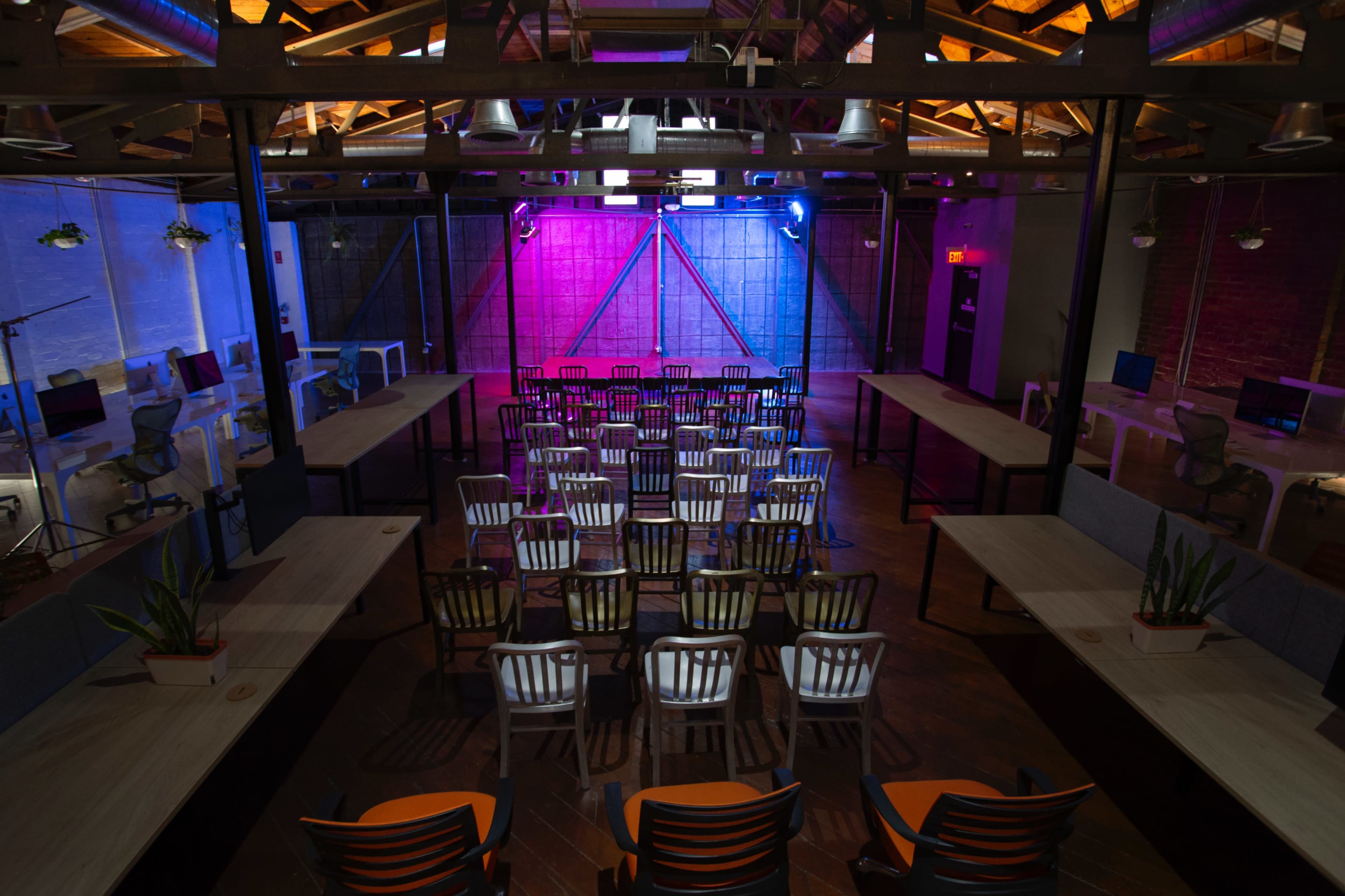 An industrial-style event space with rows of chairs facing a stage area, surrounded by tables and colorful lighting.
