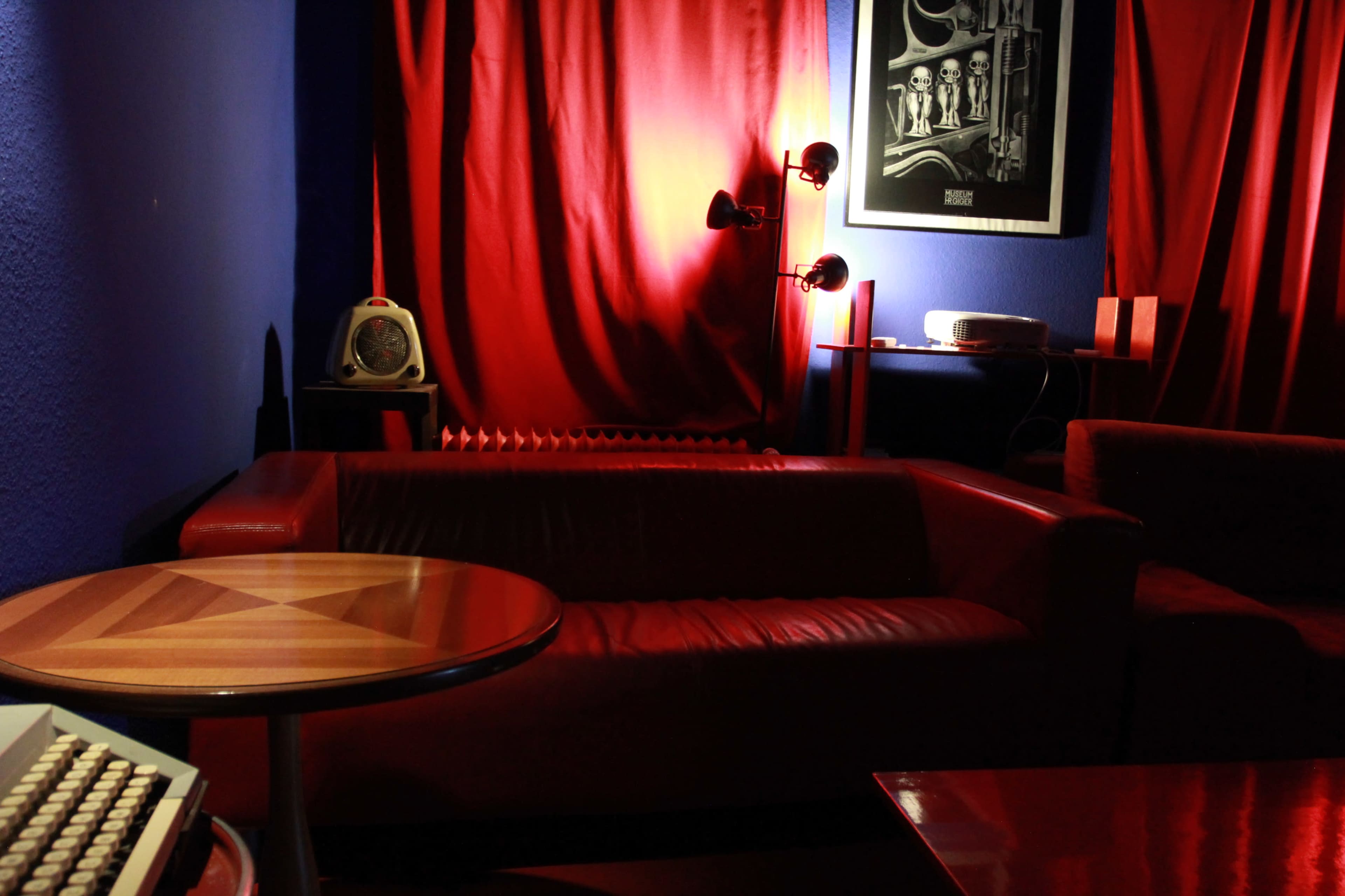 The image shows a cozy room featuring a red couch, a wooden table, and a vintage fan against a backdrop of blue walls and red curtains.