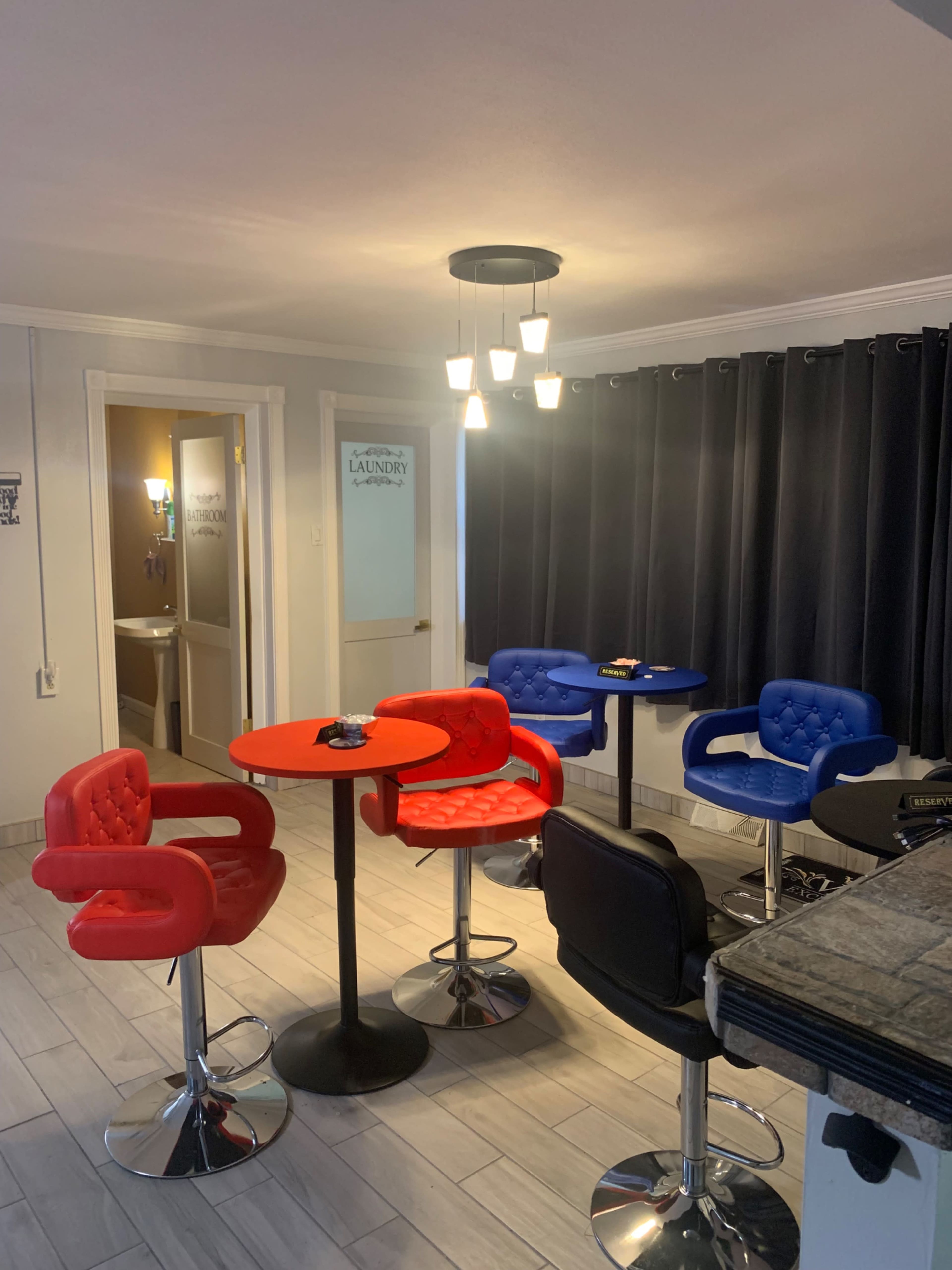 The image shows a small interior space with red and blue bar stools around black tables, accented by a hanging light fixture and large black curtains.