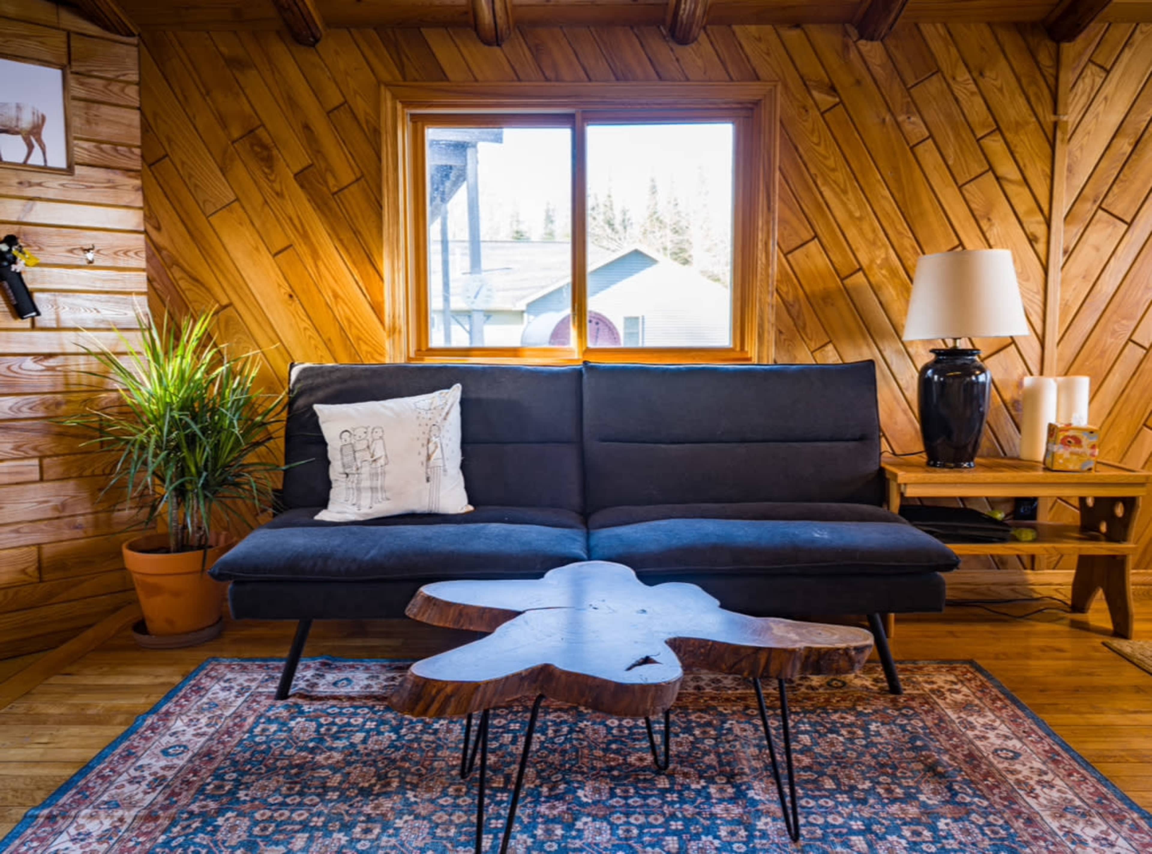 A modern living area features a dark sofa, a unique wooden coffee table, a floor lamp, and a potted plant, all set against a wooden-paneled wall and a window revealing an exterior view.
