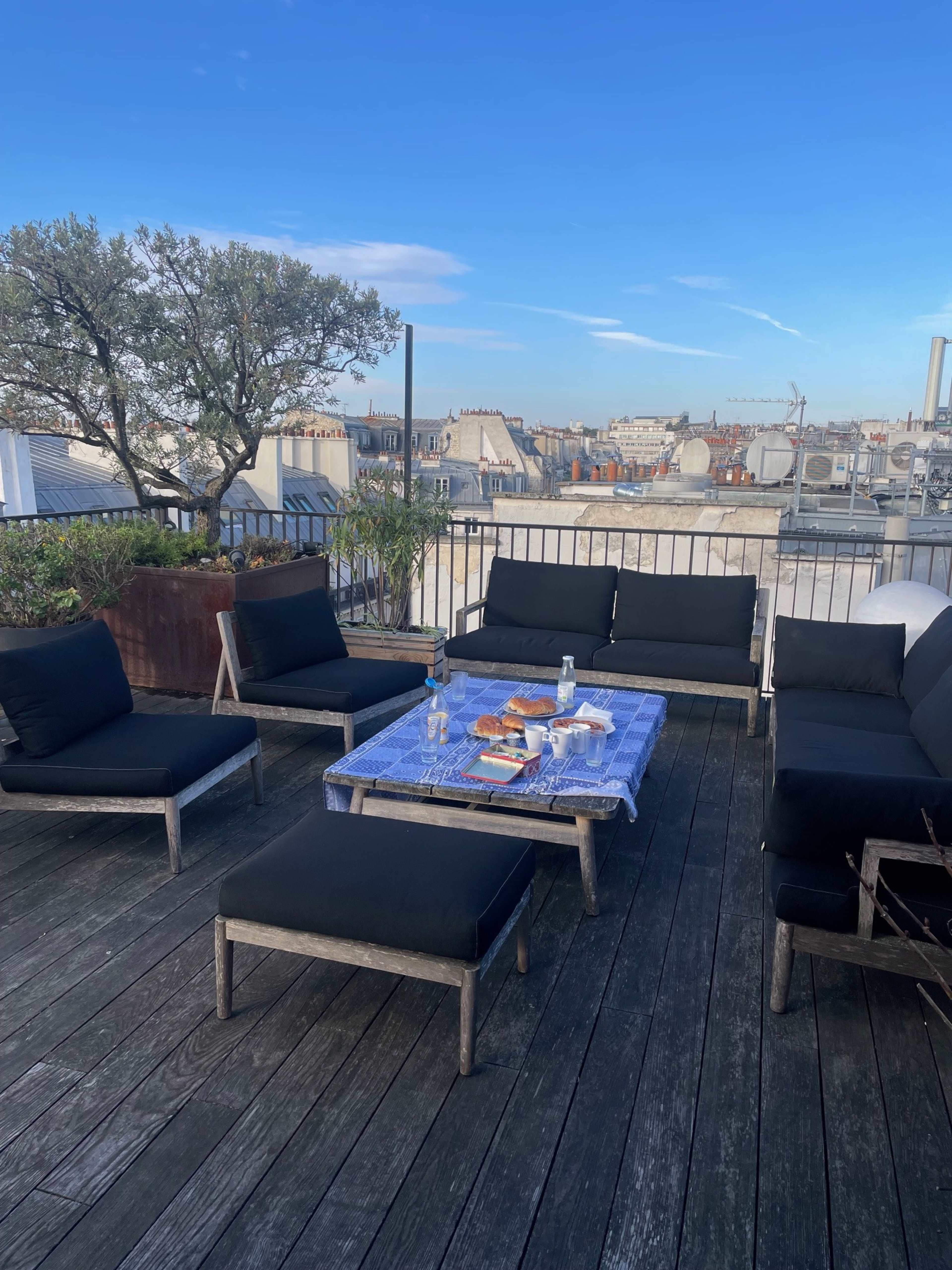 The image shows a rooftop terrace with black outdoor furniture arranged around a table, overlooking a cityscape under a clear blue sky.