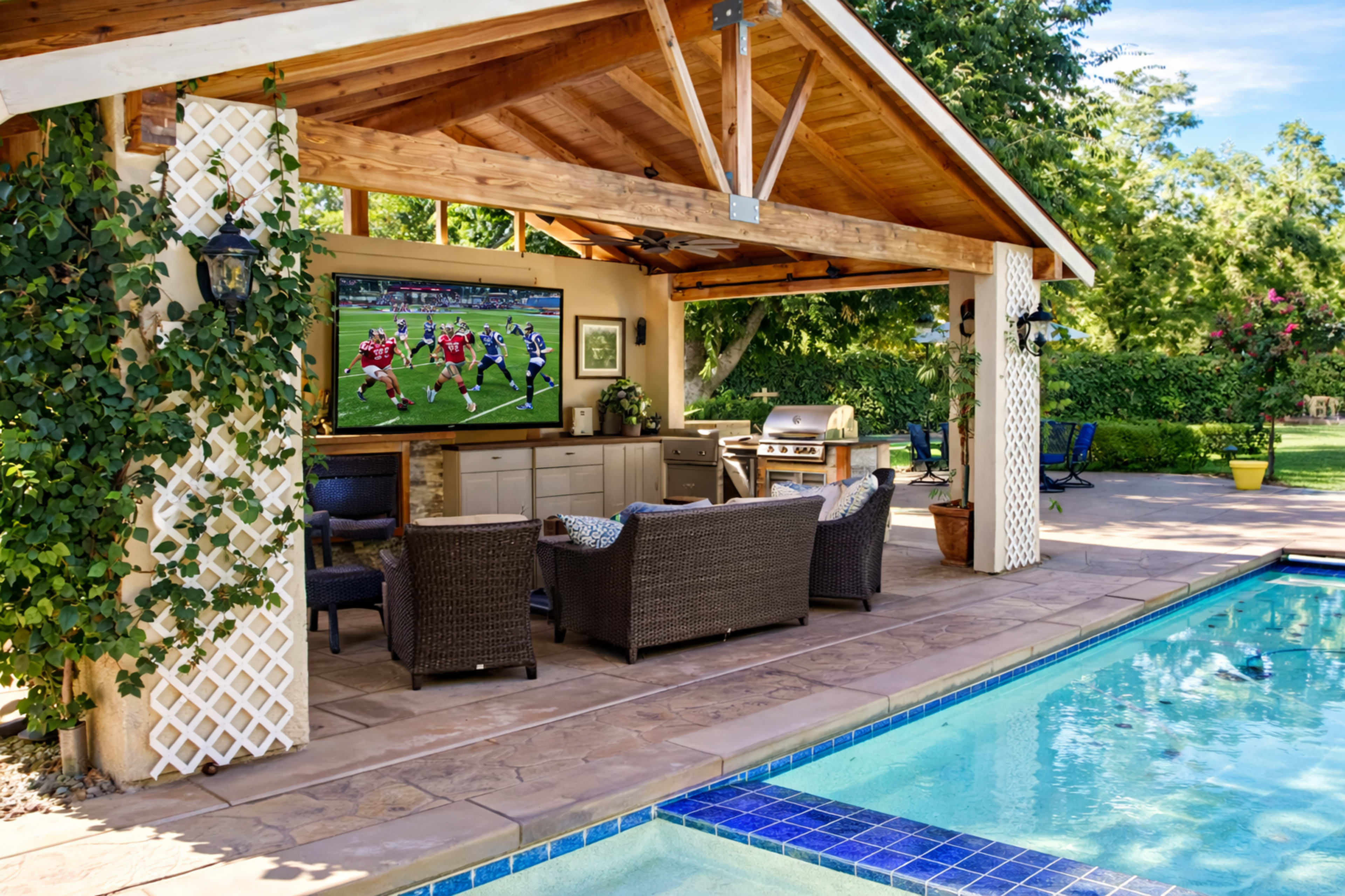 A covered outdoor patio with a television, seating area, and a barbecue grill overlooks a swimming pool surrounded by green landscaping.