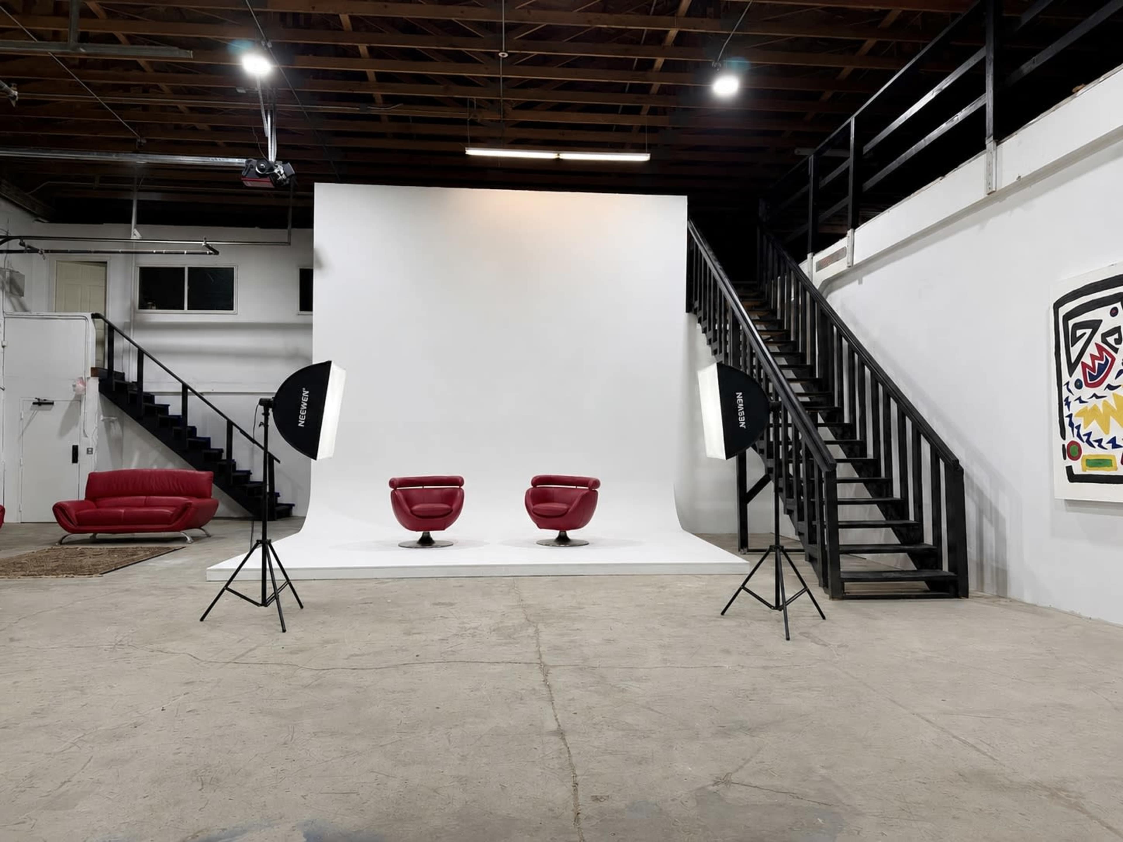 The image shows a modern studio space featuring a white backdrop, two red chairs, and lighting equipment, with a staircase and artwork visible in the background.