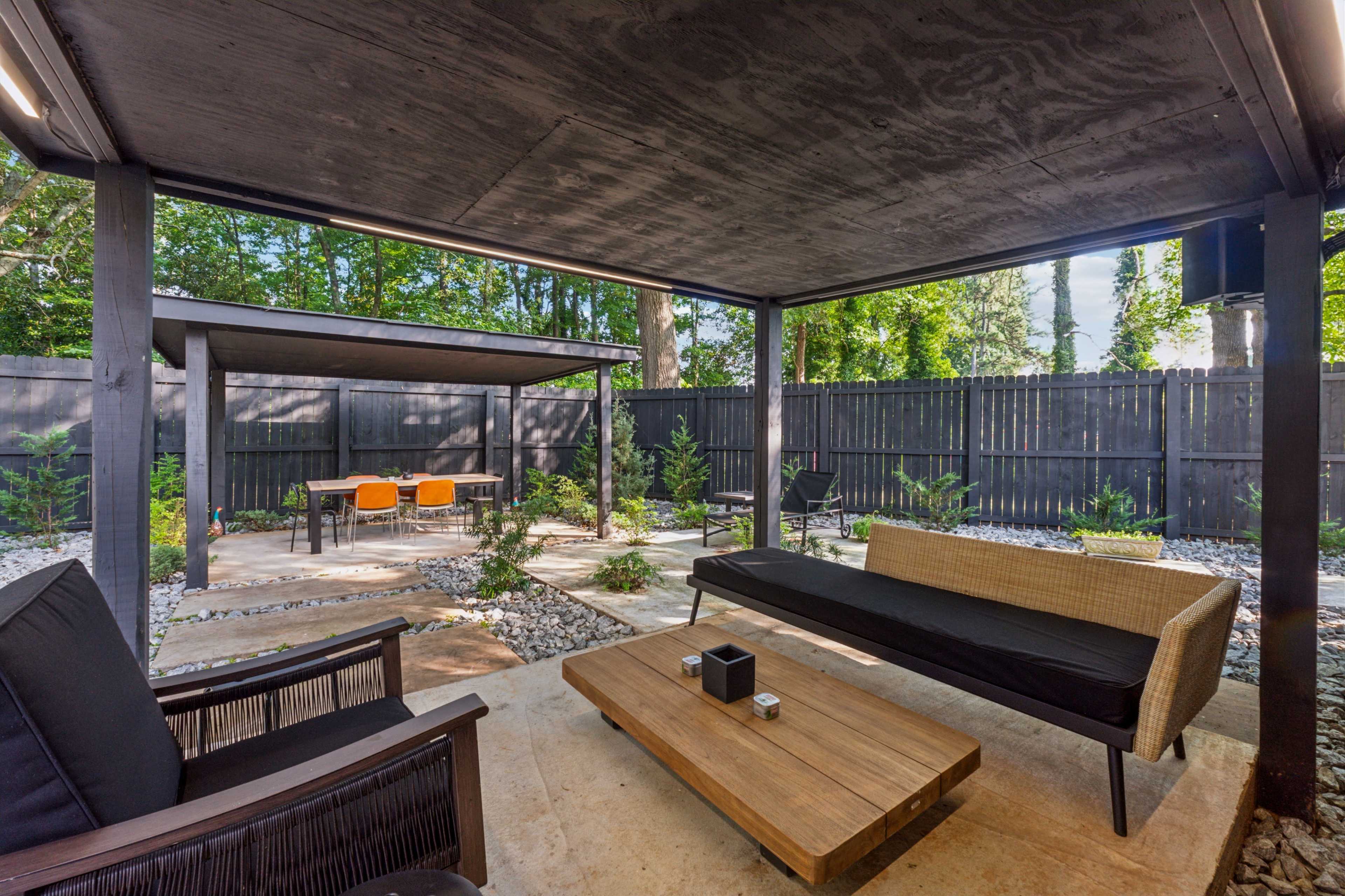 The image shows a covered patio area with modern outdoor seating, a wooden coffee table, and a garden featuring gravel pathways and small plants.
