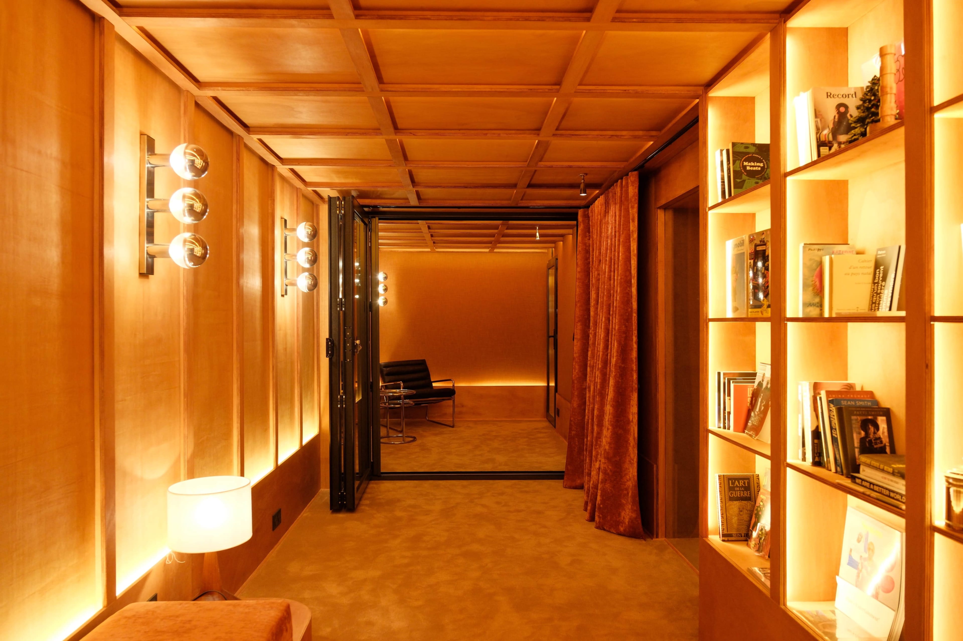 The image shows a well-lit hallway with wood paneling, featuring bookshelves on one side and soft, contemporary furniture along with a curtain on the other.