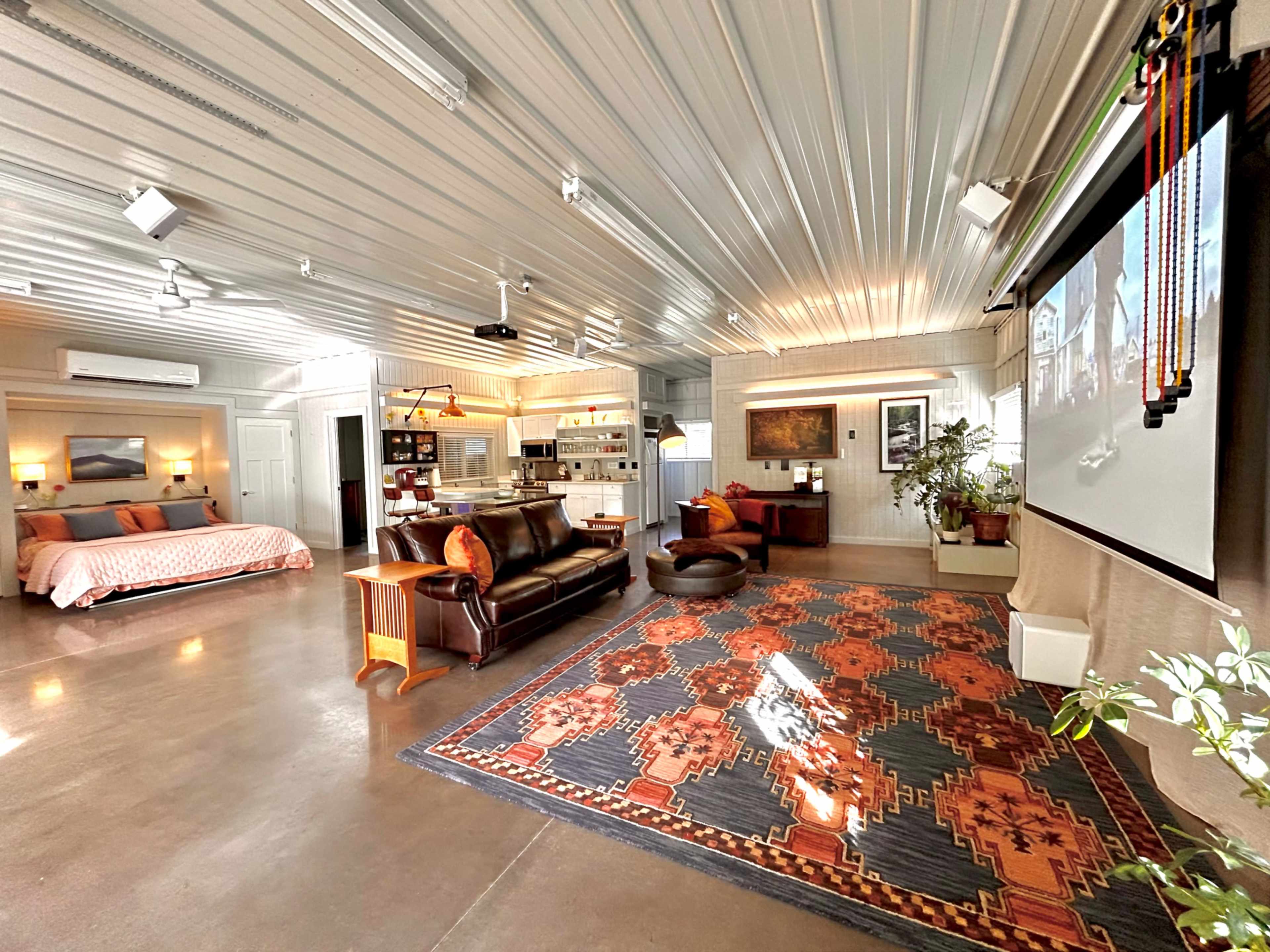 The image shows a spacious, modern living area featuring a large bed, seating arrangements, a kitchen area, and a projection screen on one wall.