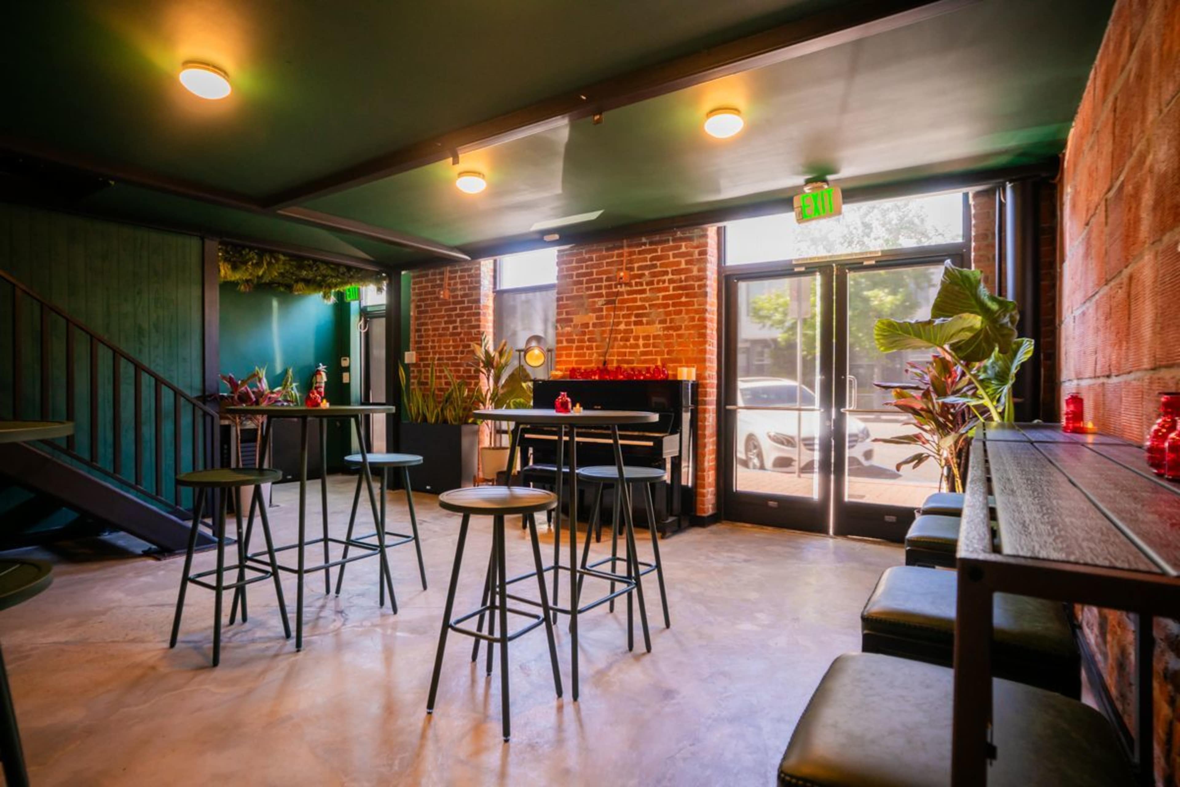 The image shows a modern bar area with metal high-top tables, a brick wall, and green plants, featuring an entrance with large glass doors.