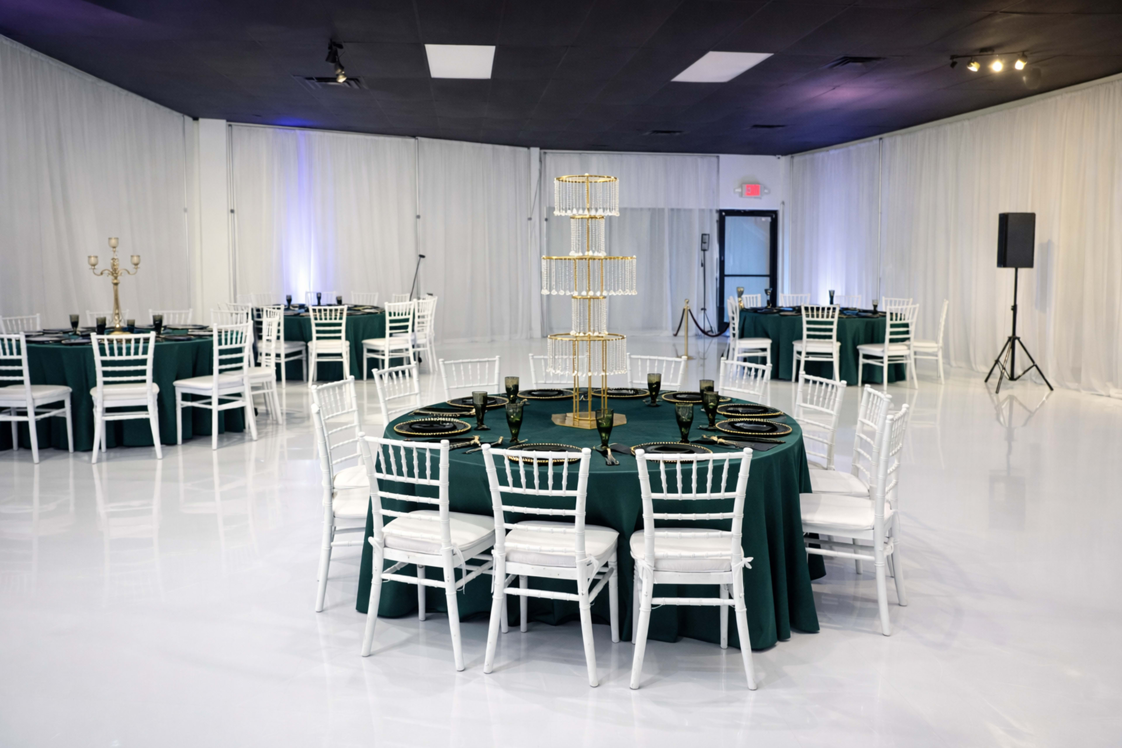 The image depicts an elegantly arranged indoor event space featuring round tables covered in dark green tablecloths, white chairs, and a tiered serving stand at the center.