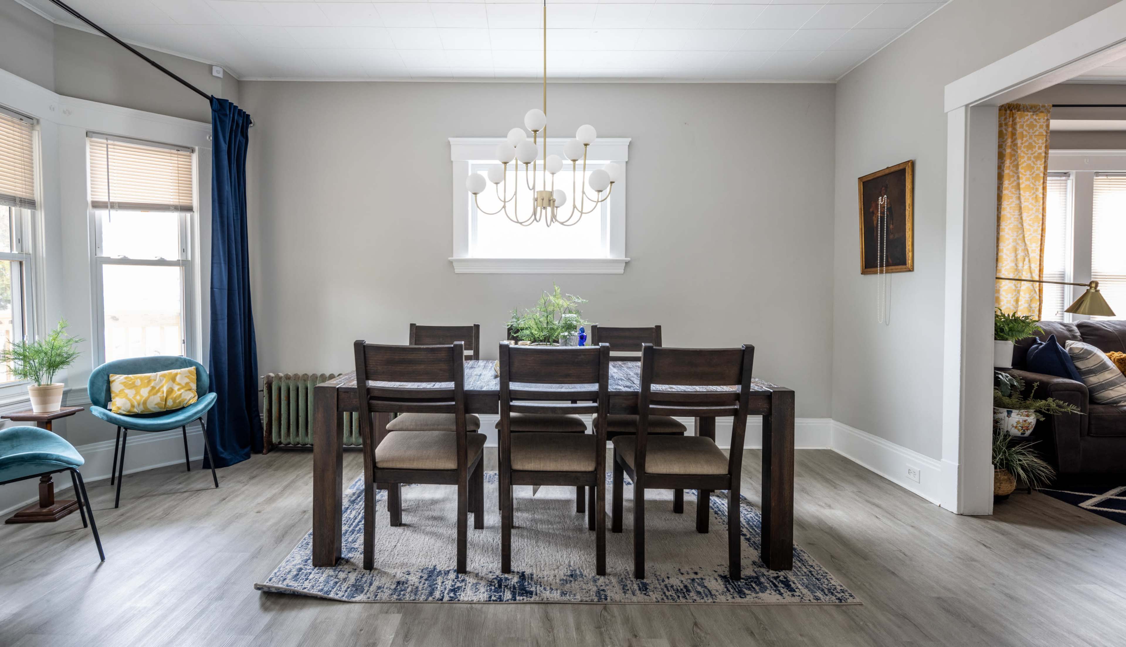 A dining room features a long wooden table with six chairs, a chandelier overhead, and windows allowing natural light to enter.