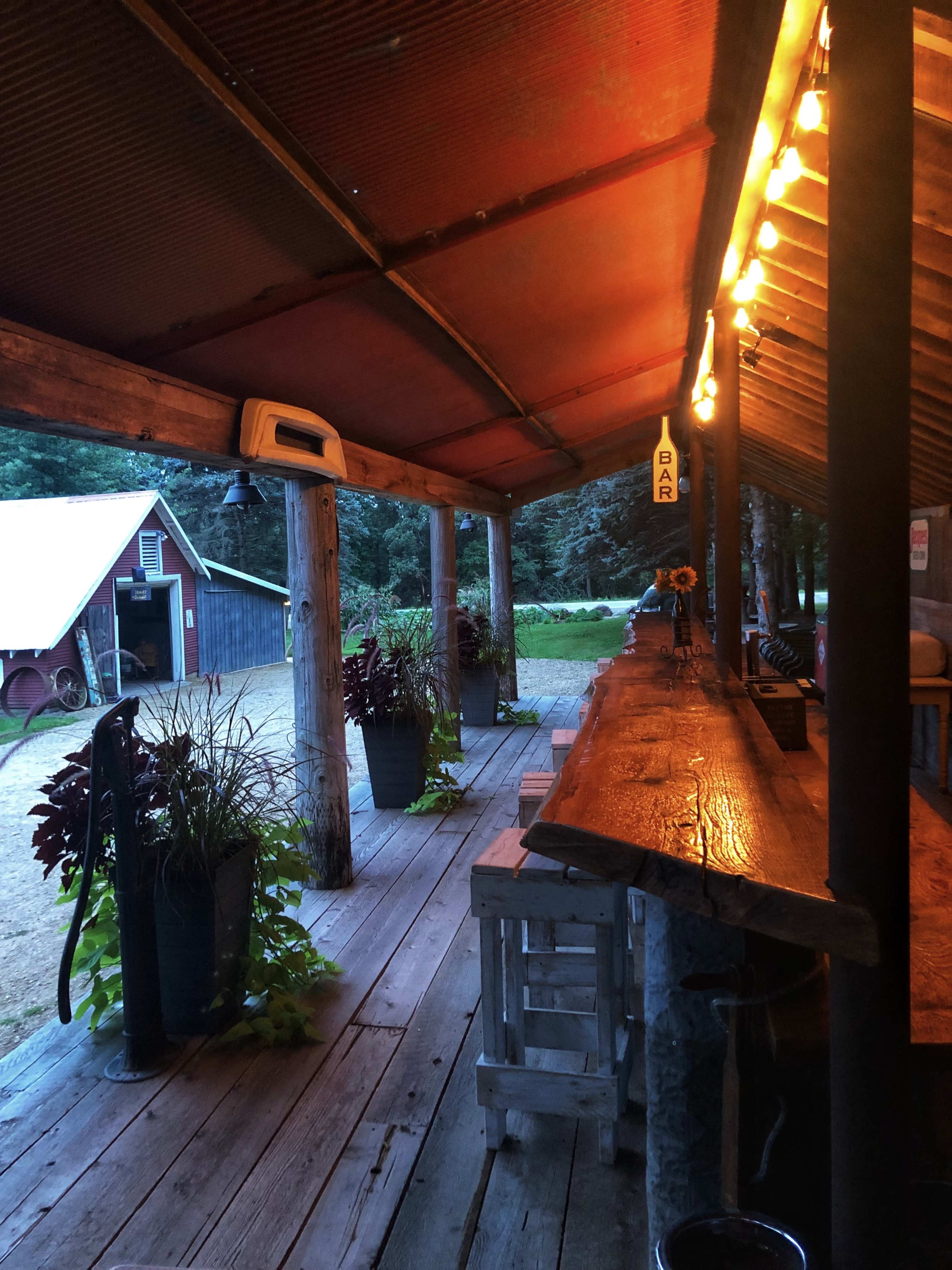 The image shows a rustic outdoor bar area with wooden benches and a long tabletop, illuminated by string lights, alongside a green lawn and a red barn in the background.