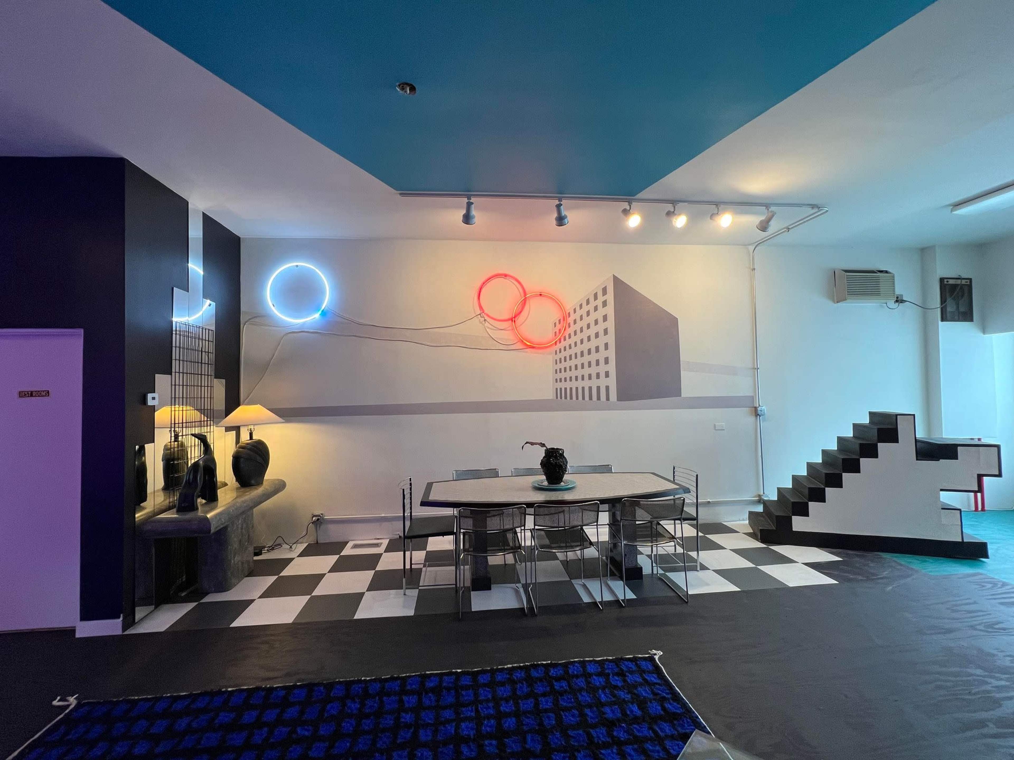 The image shows a modern interior space featuring a dining area with a black and white checkered floor, a table and chairs, and colorful neon wall art.
