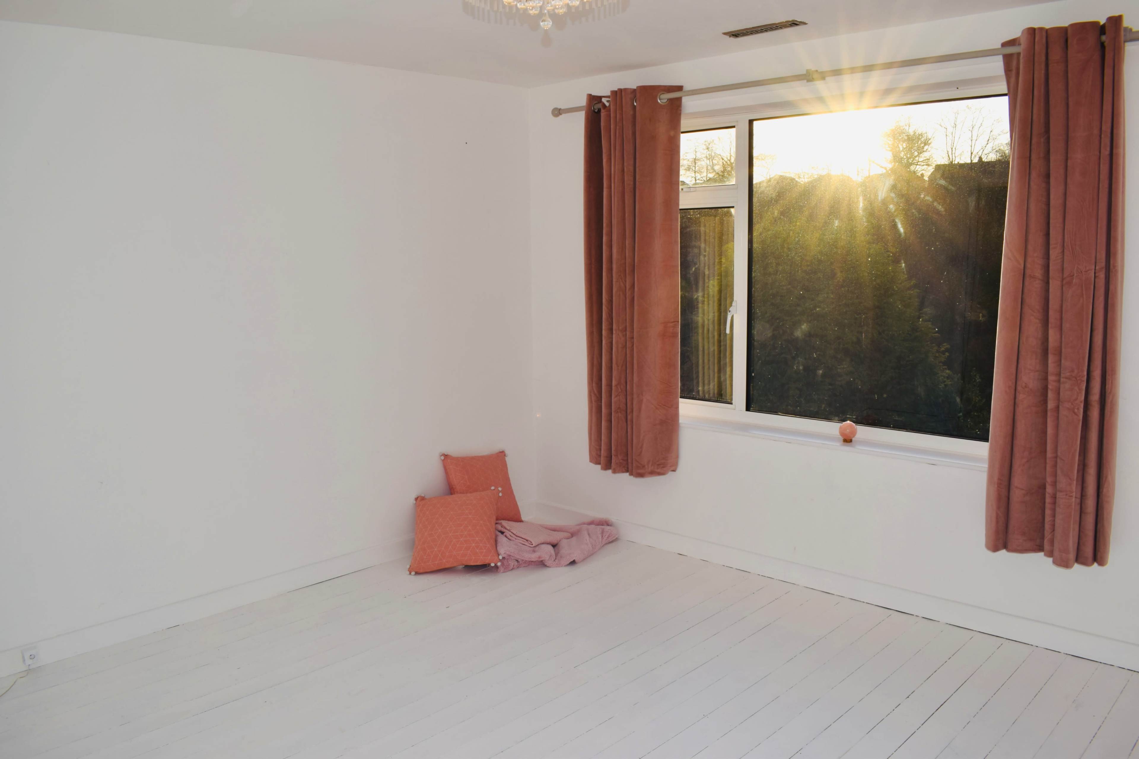 A bright, empty room with white walls and a large window adorned with rust-colored curtains, featuring a small pillow and blanket on the floor.