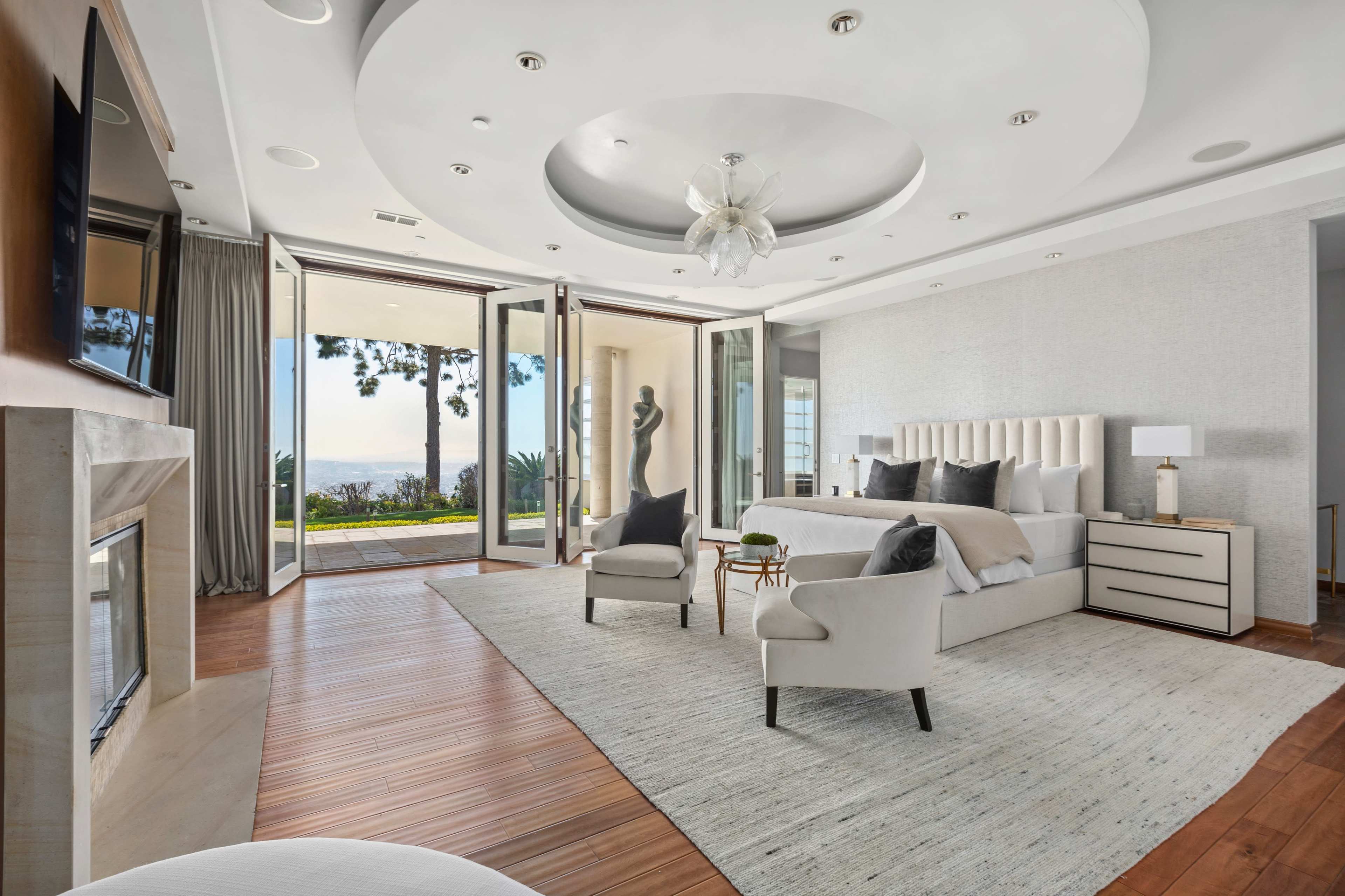 The spacious bedroom features a king-sized bed, two armchairs, a fireplace, and large sliding glass doors that open to a garden view.