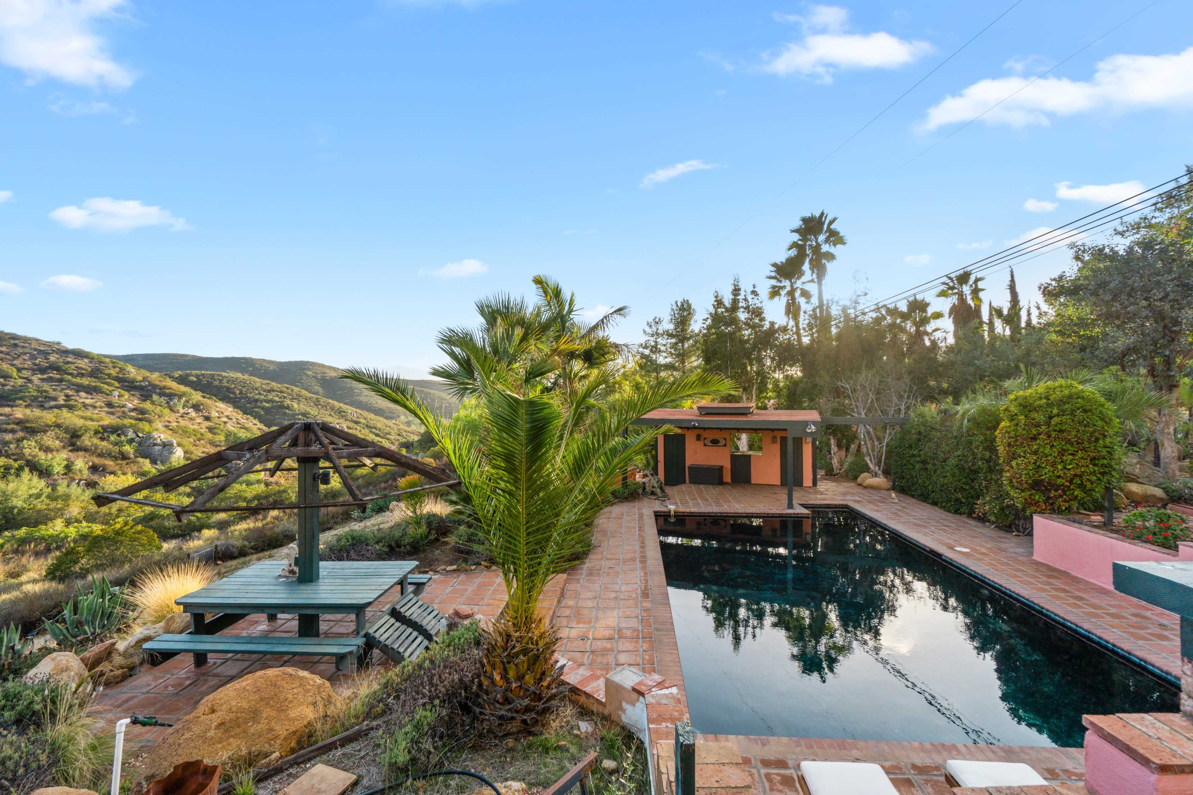 The image shows a rectangular swimming pool surrounded by palm trees, a gazebo, and a wooden deck, set against a backdrop of hills and clear blue skies.