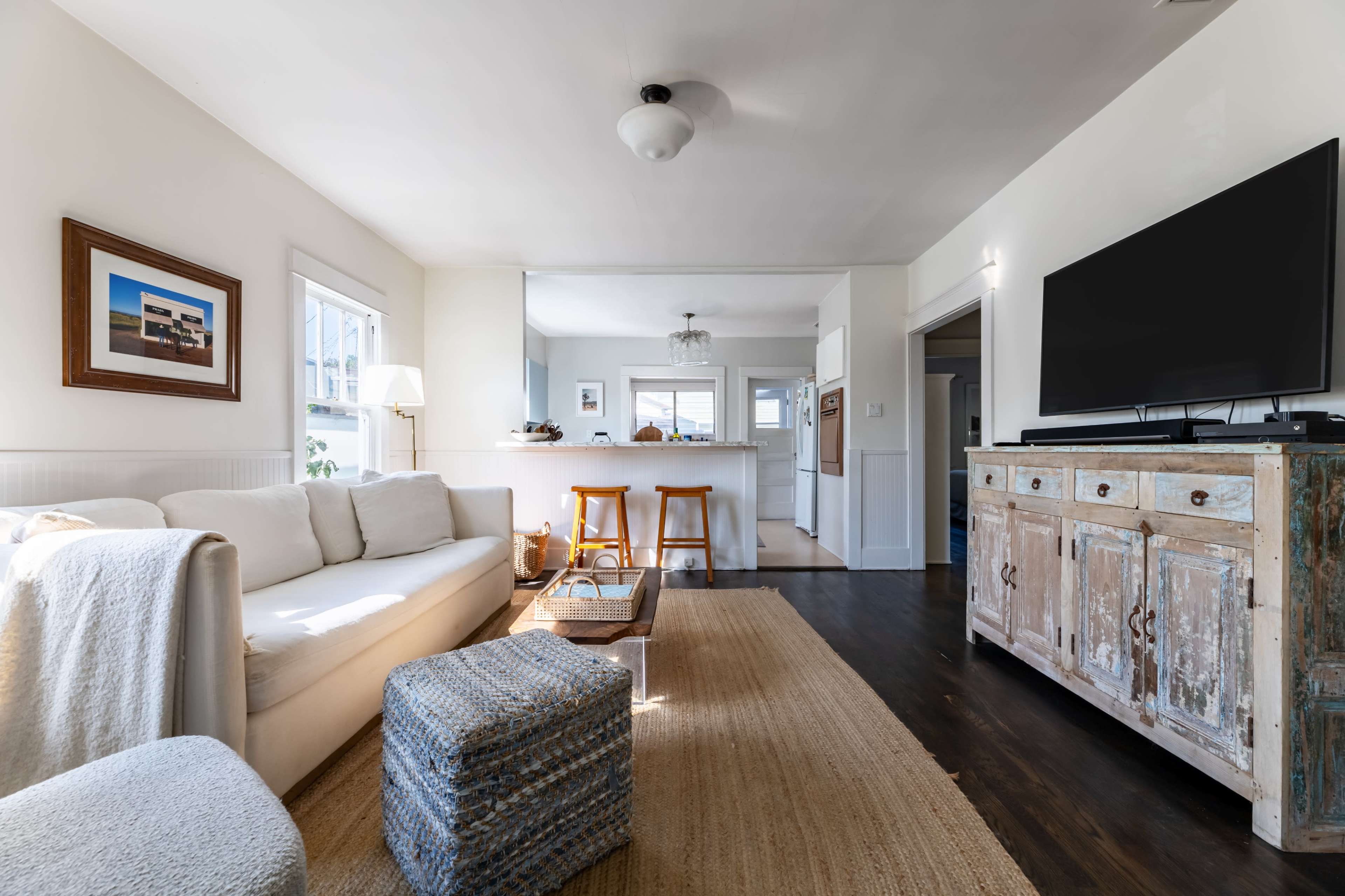 A bright, modern living room with a white sofa, wooden furniture, and a view into a kitchen area.