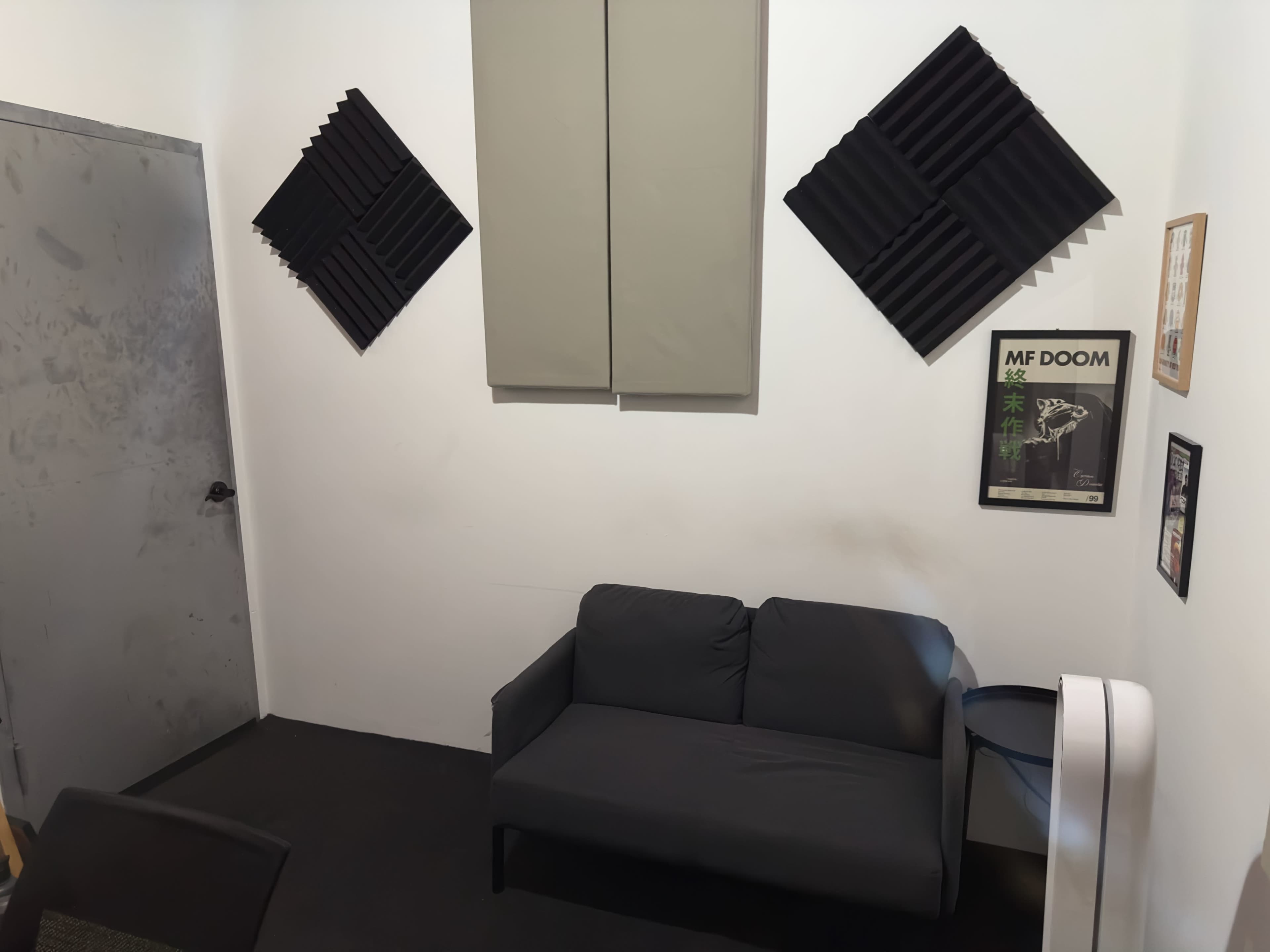 The image shows a small, simple room with a gray couch, soundproofing panels on the walls, and a framed poster beside a door.