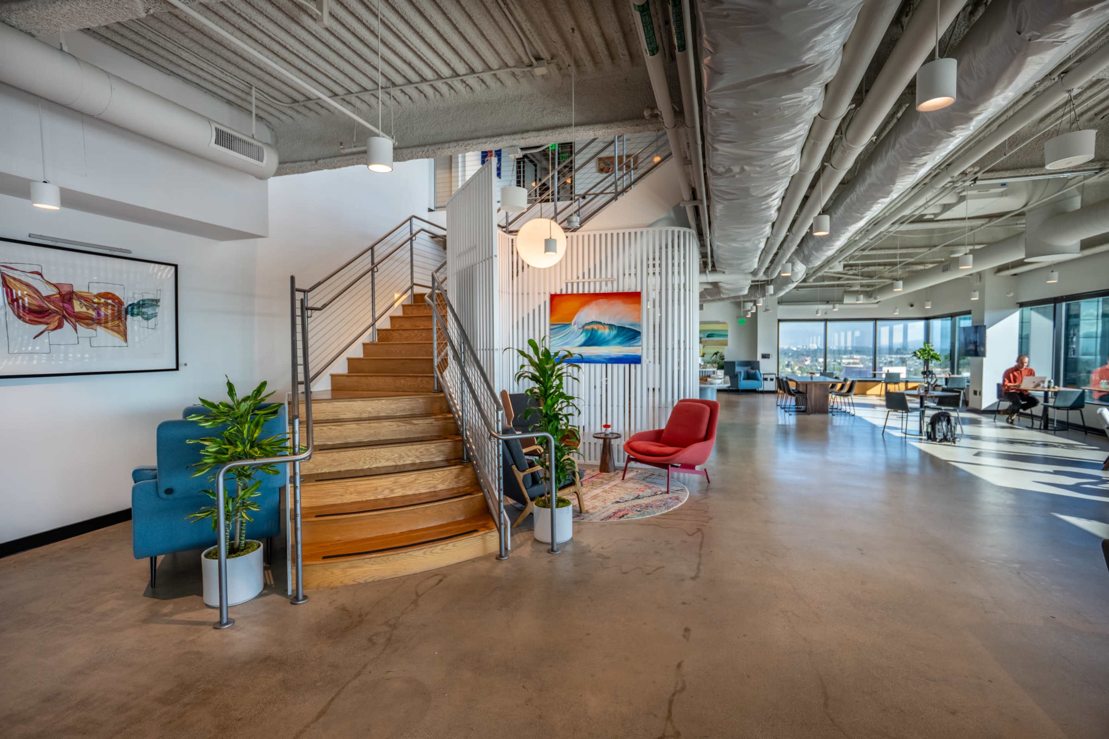 The image shows a modern office space with a staircase leading to an upper level, surrounded by plants, artwork, and seating areas.