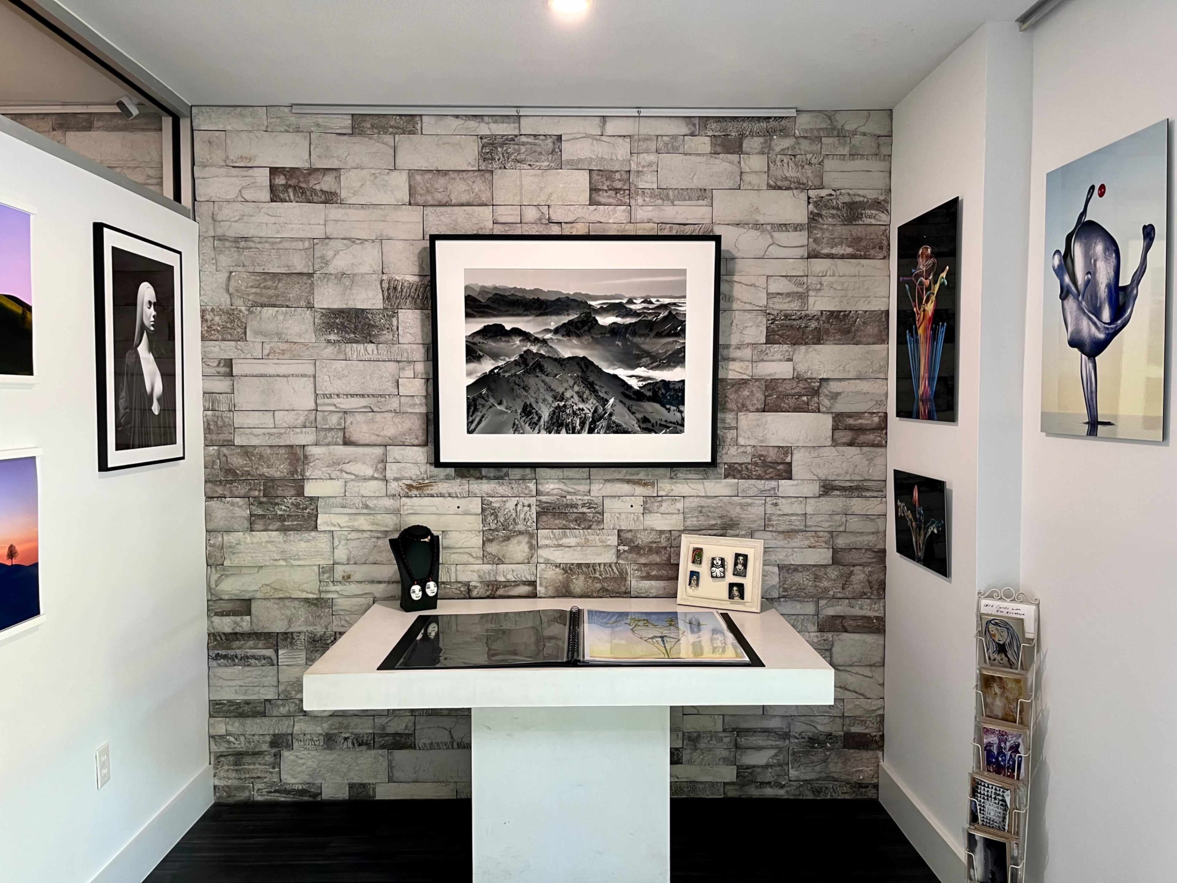 A modern gallery space with a stone wall, featuring framed photographs and artwork, and a central display table with two framed pictures and a black object on it.