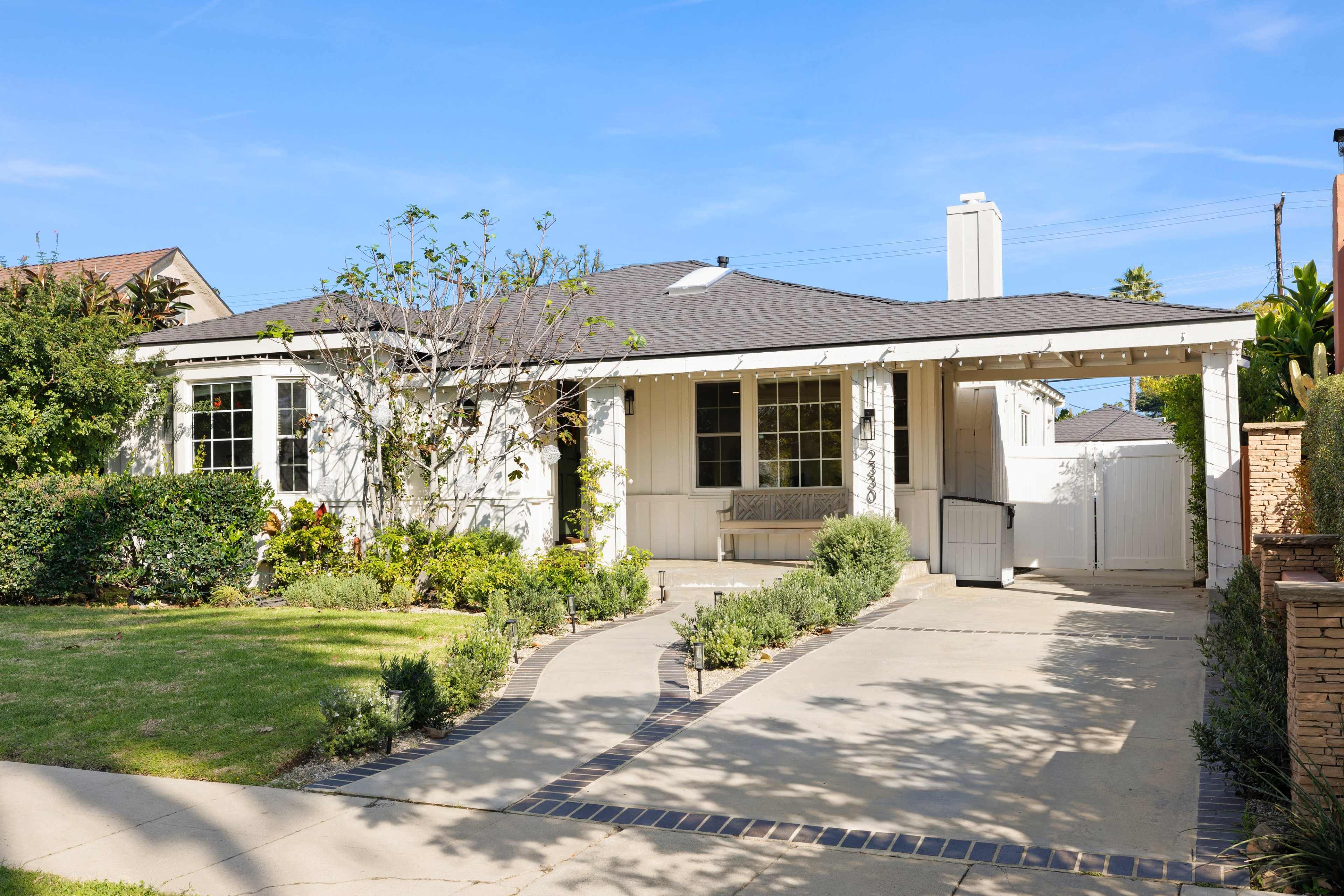 The image shows a single-story house with a front yard featuring shrubs and a pathway leading to a covered porch.