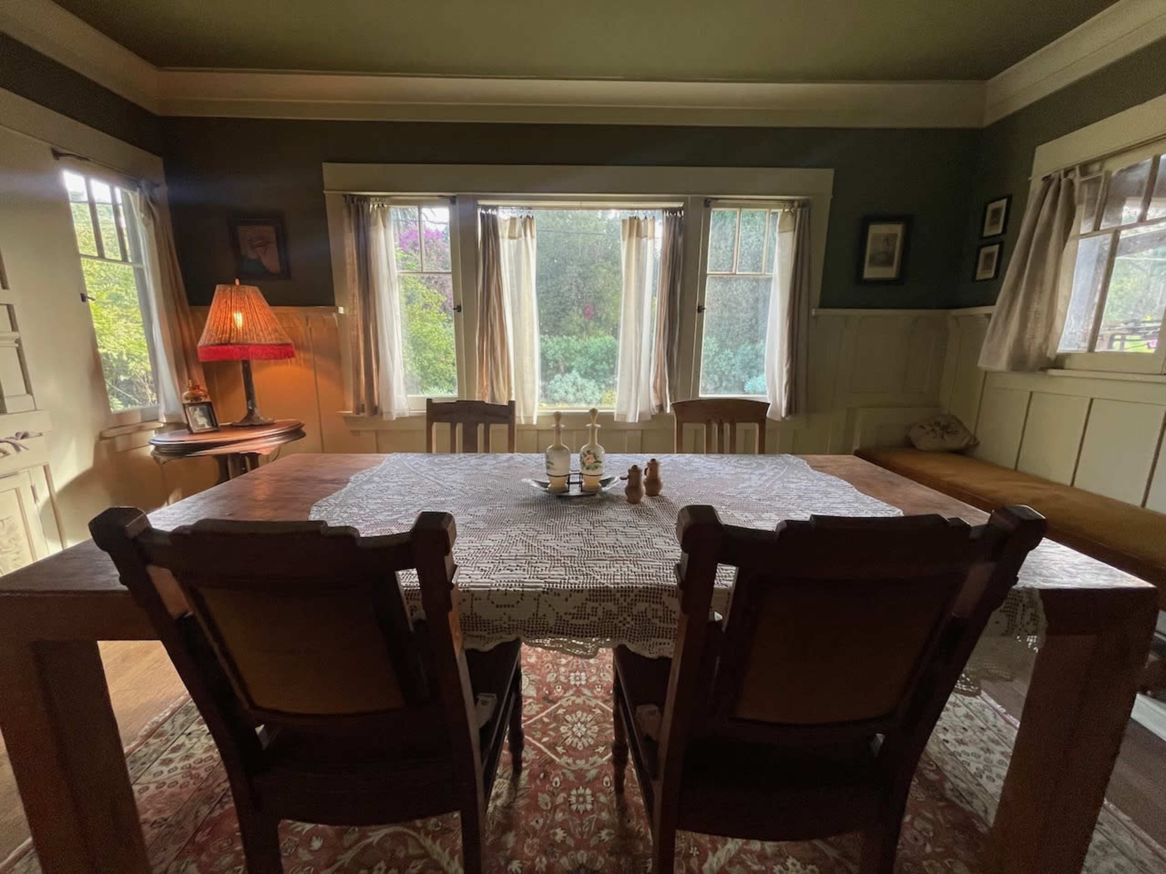 A wooden dining table is set with a decorative tablecloth, flanked by two chairs and illuminated by natural light coming through large windows.