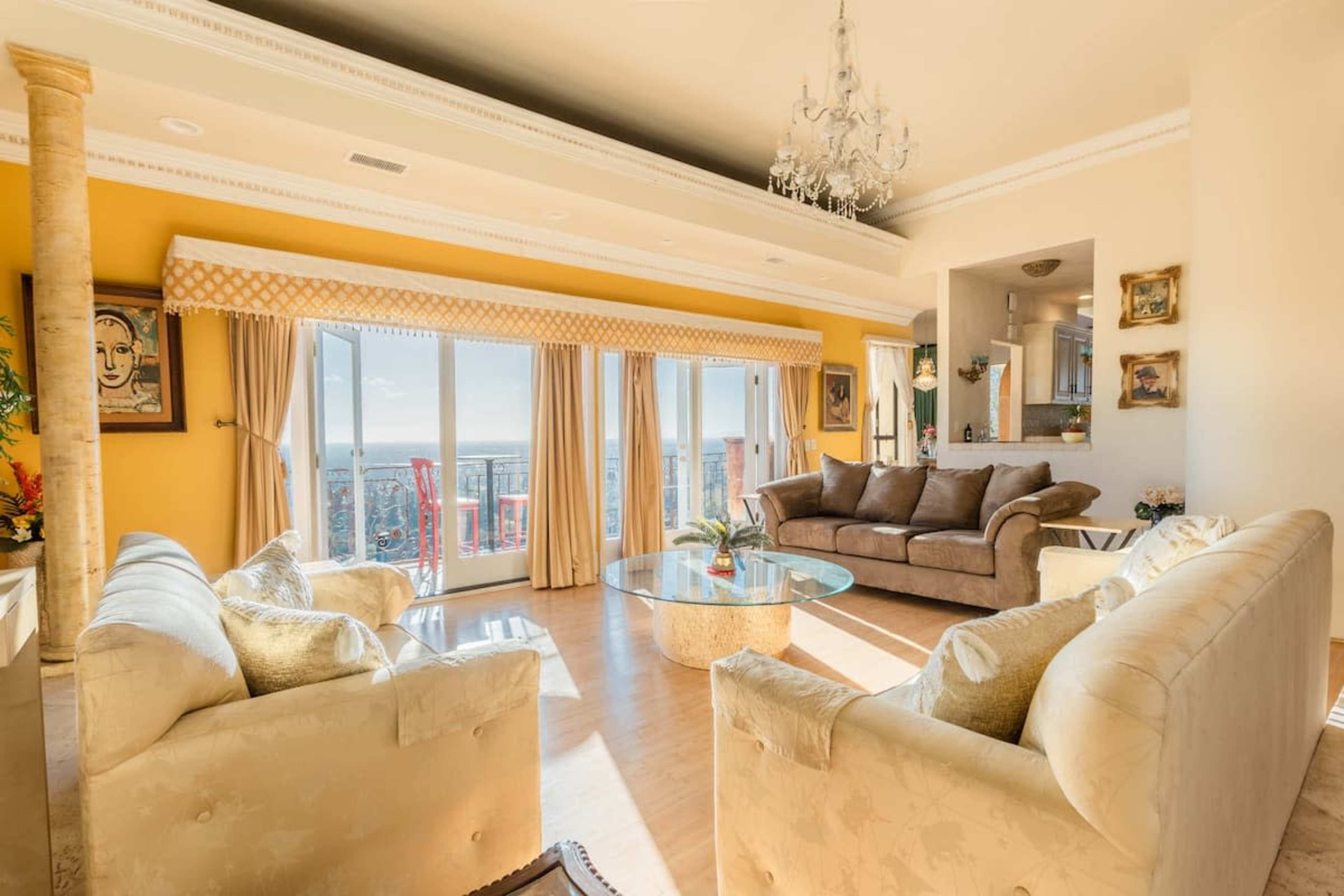 A brightly lit living room with a chandelier, two plush sofas, a glass coffee table, and large windows overlooking a scenic view.