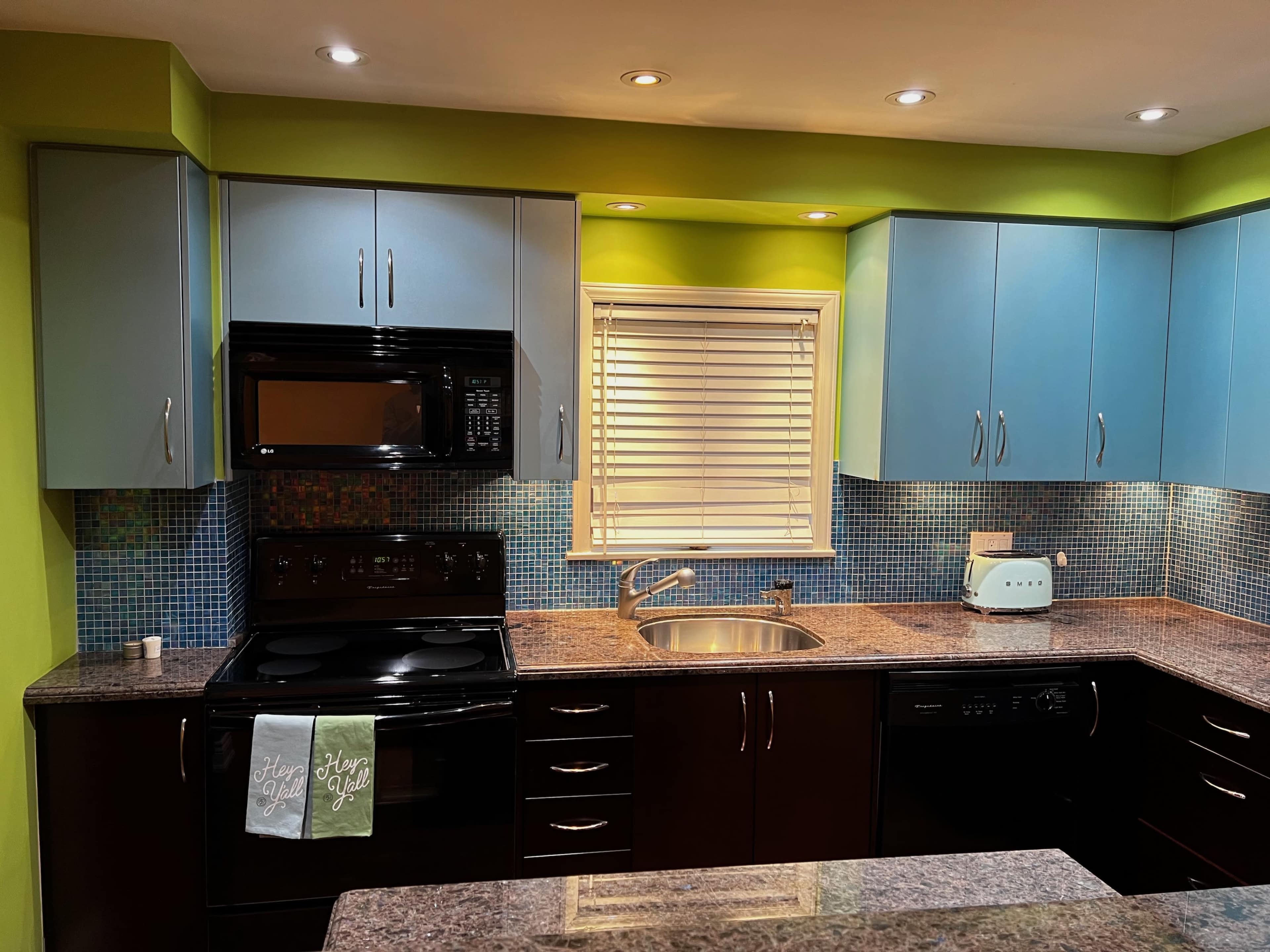 A modern kitchen with blue upper cabinets, dark lower cabinets, and a green accent wall, featuring a sink, black appliances, and a tiled backsplash.
