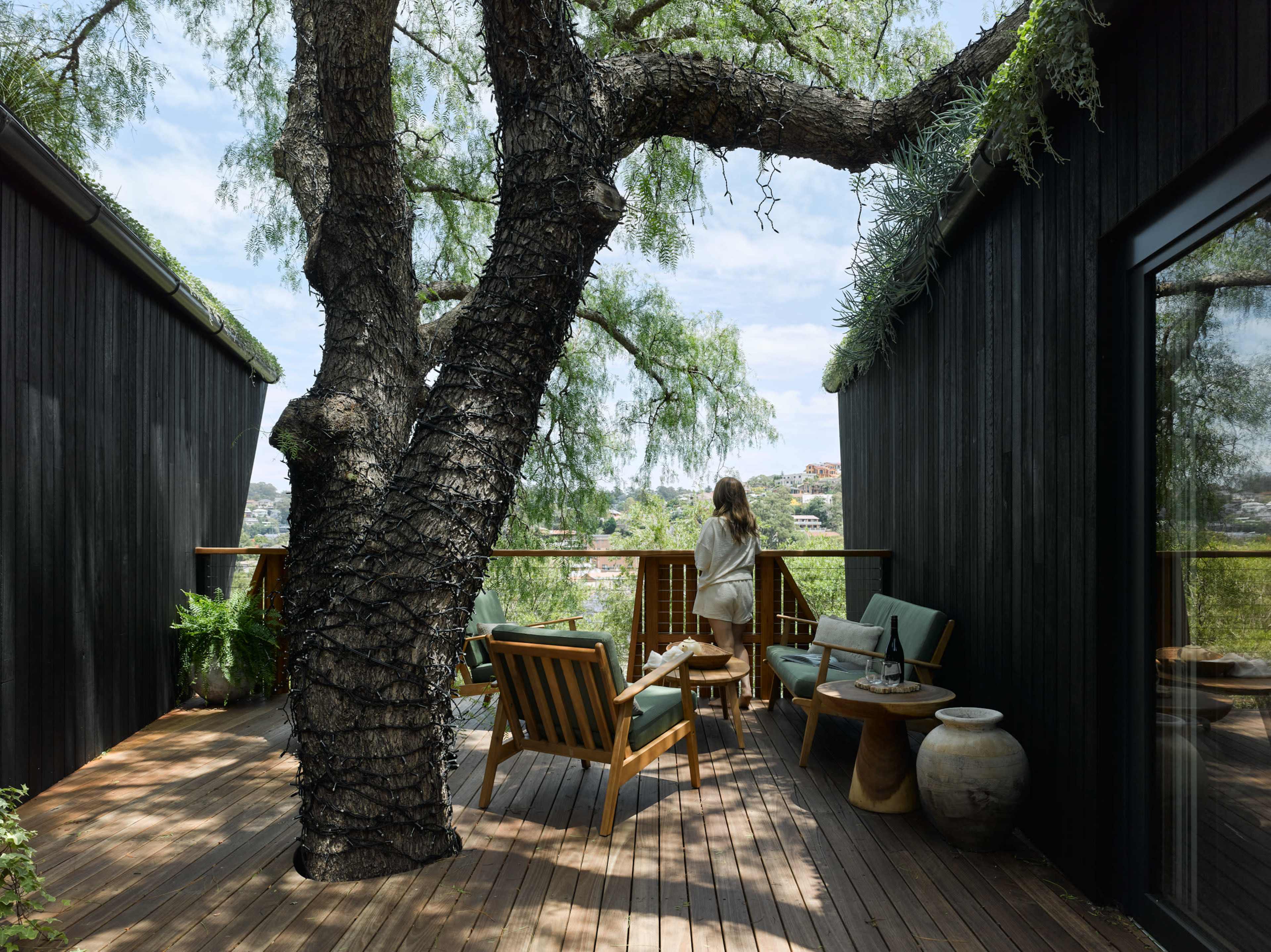 A woman stands on a wooden deck overlooking a landscape, surrounded by a large tree and modern furnishings.