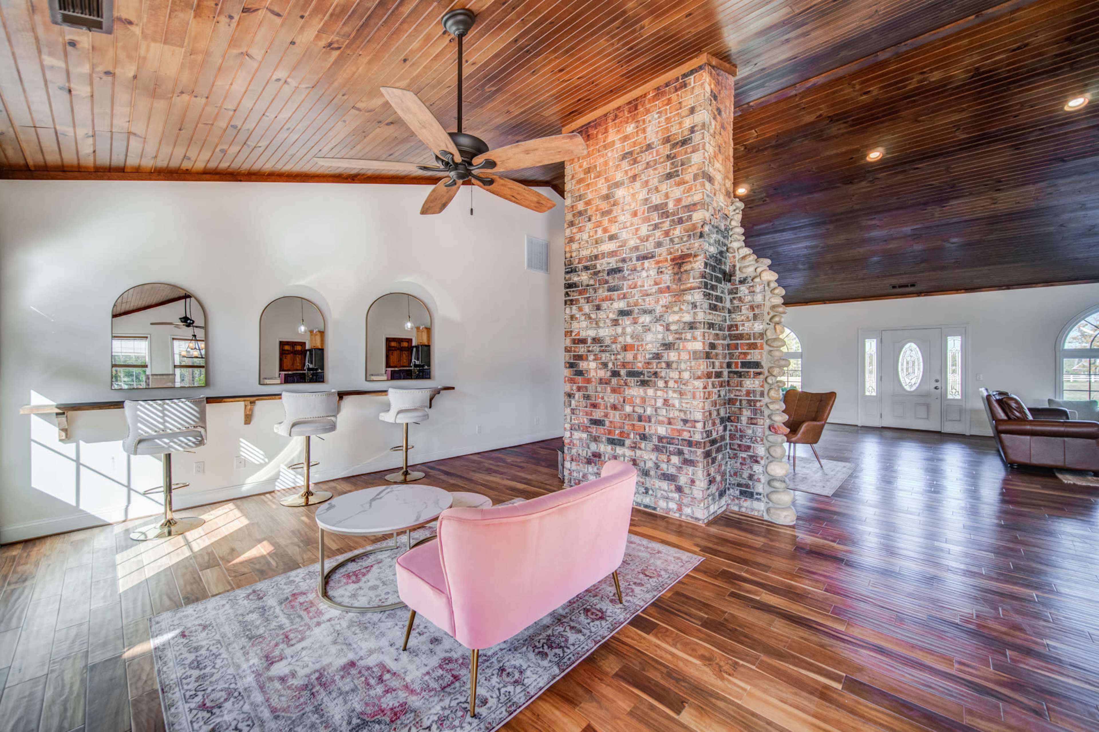 The image shows a living space with a wooden ceiling, a brick feature wall, and modern furnishings including a pink chair and a bar area.