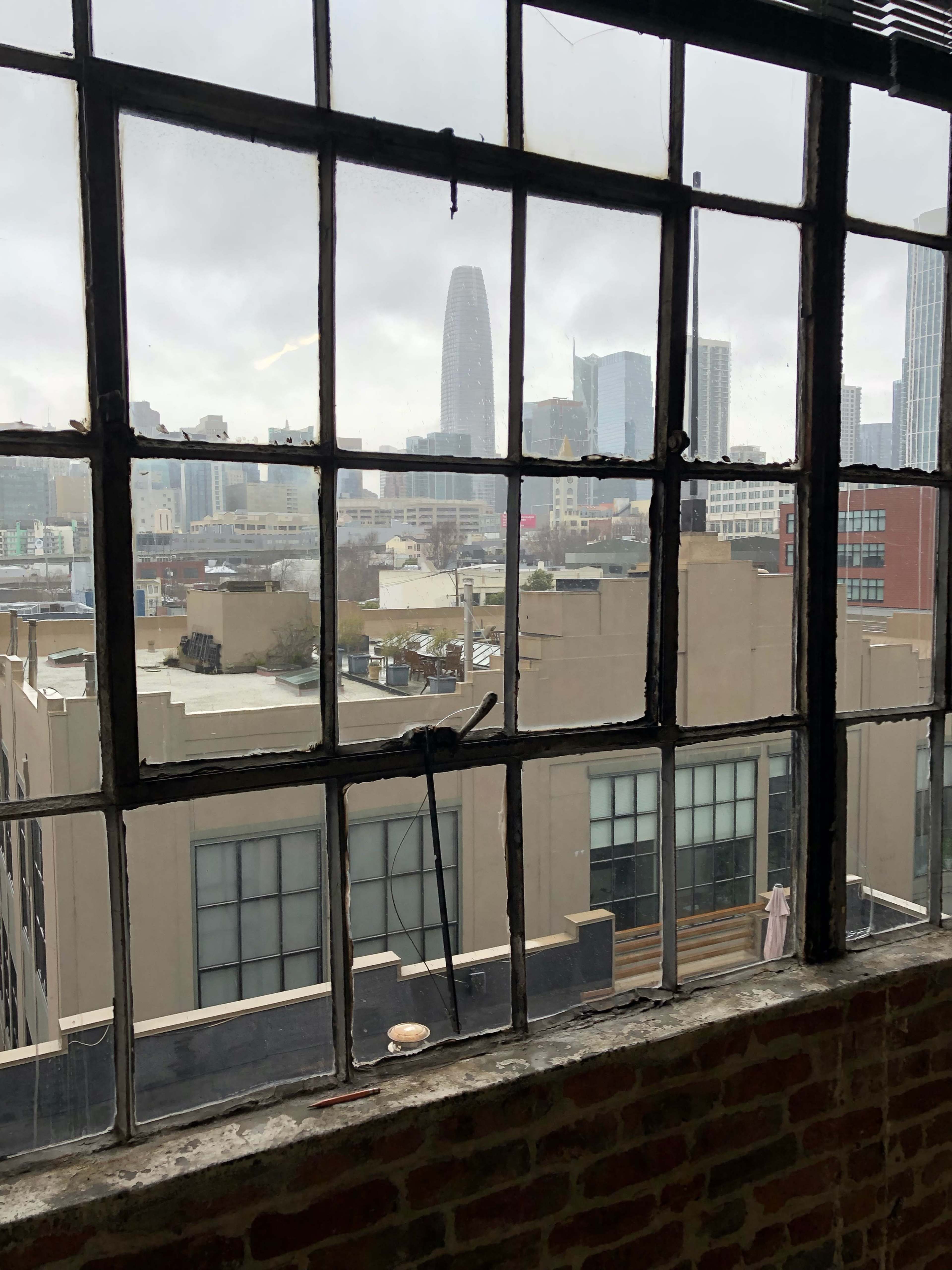 A large industrial window frame with multiple panes, through which a city skyline with tall buildings is visible on a cloudy day.