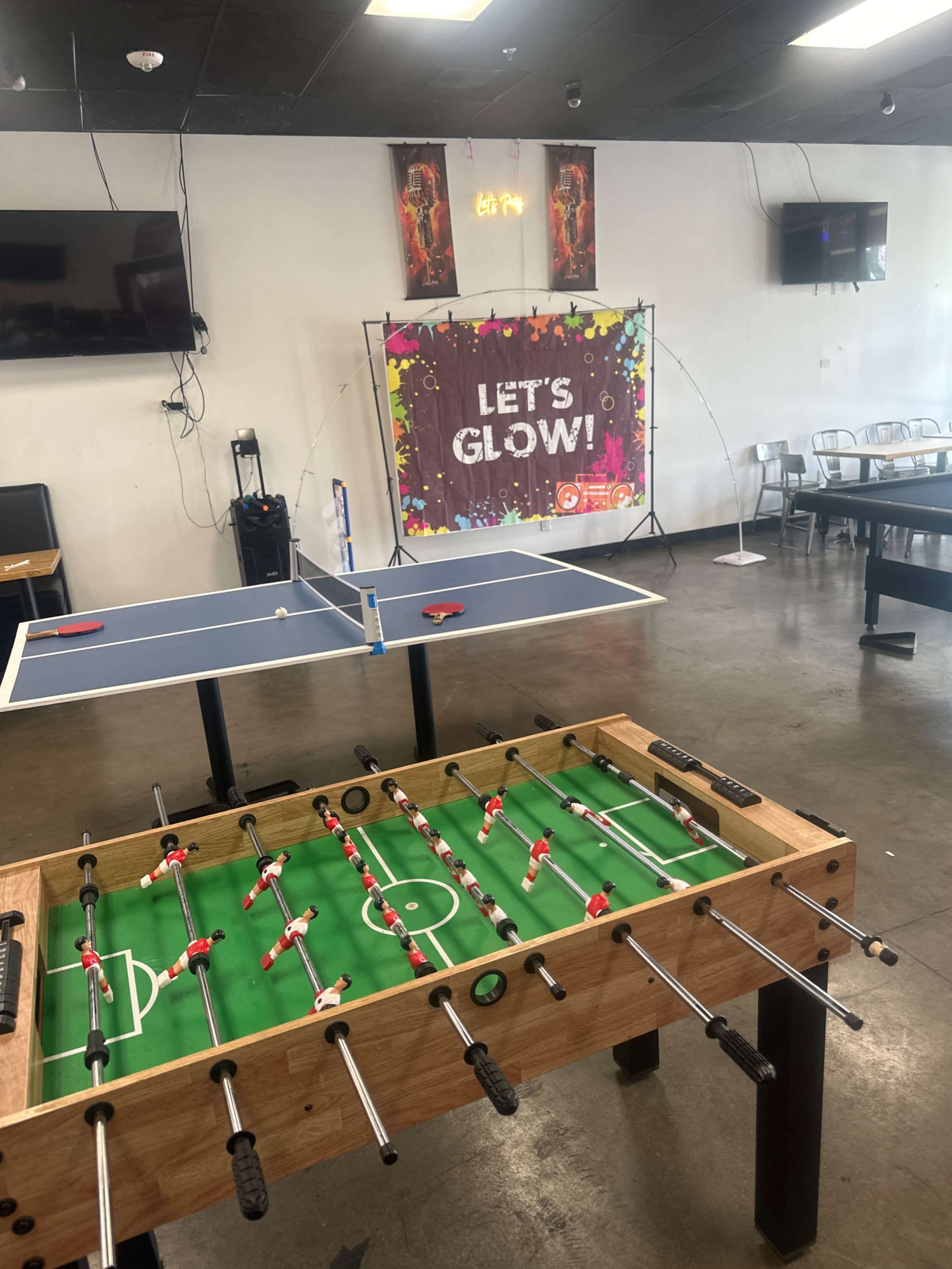 The image shows a recreation area with a foosball table in the foreground, a ping pong table in the background, and a colorful "LET'S GLOW!" banner on the wall.