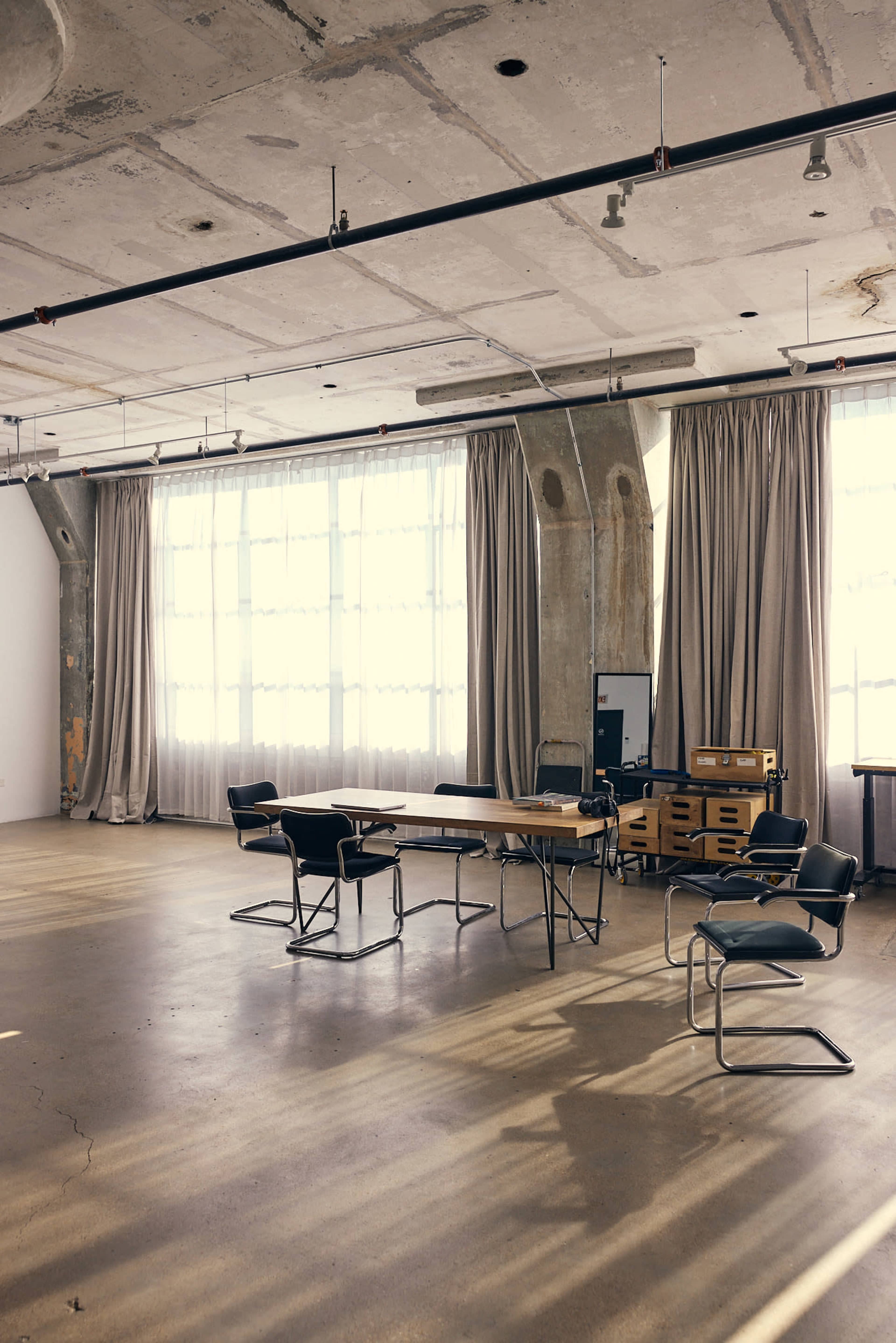 A minimalist meeting room features a rectangular table surrounded by several chairs, with large windows letting in natural light and curtains partially drawn.