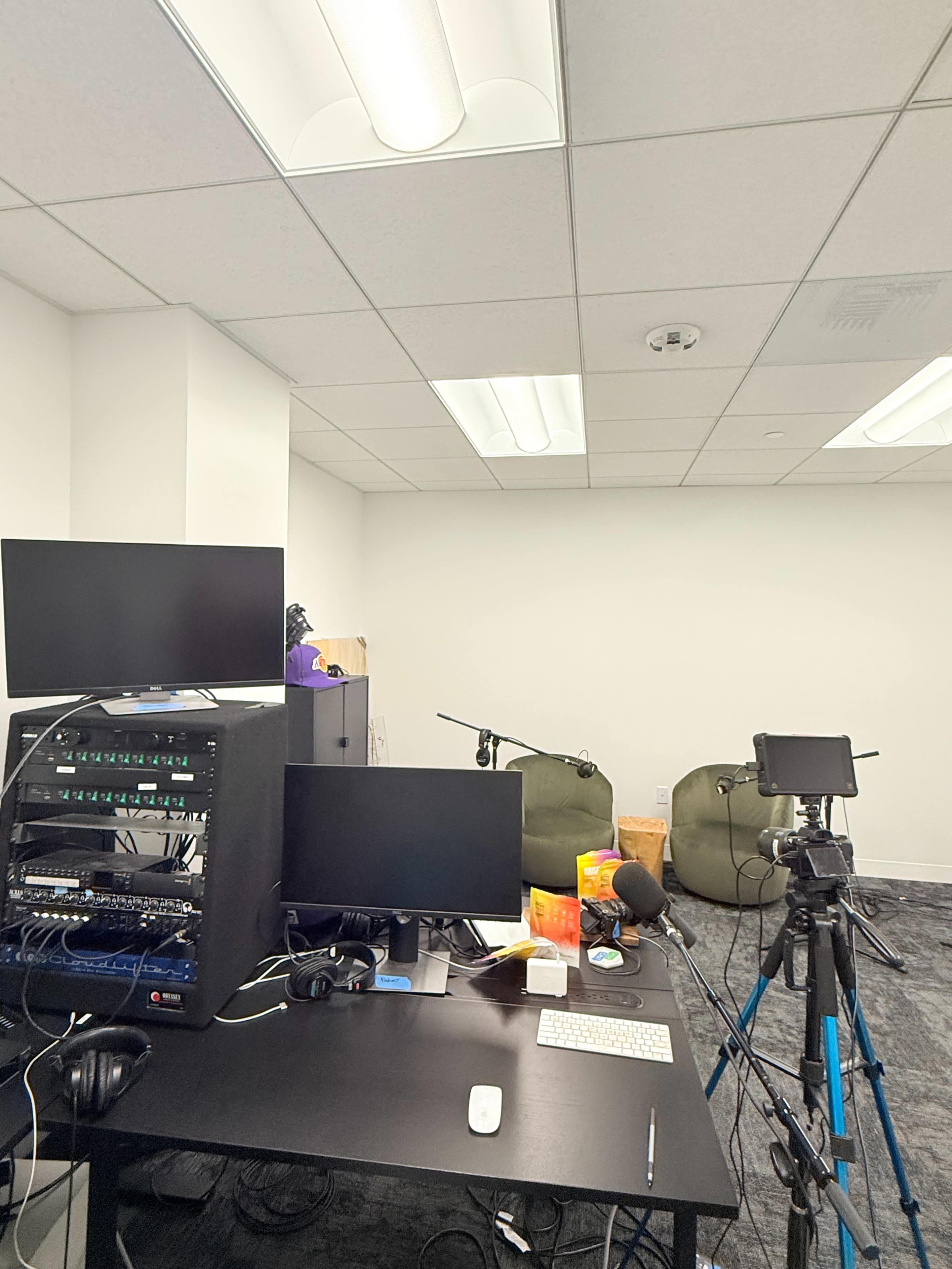 The image shows a minimal office space with two computers on a dark desk, a rack of audio equipment, and two large green bean bags in the background.