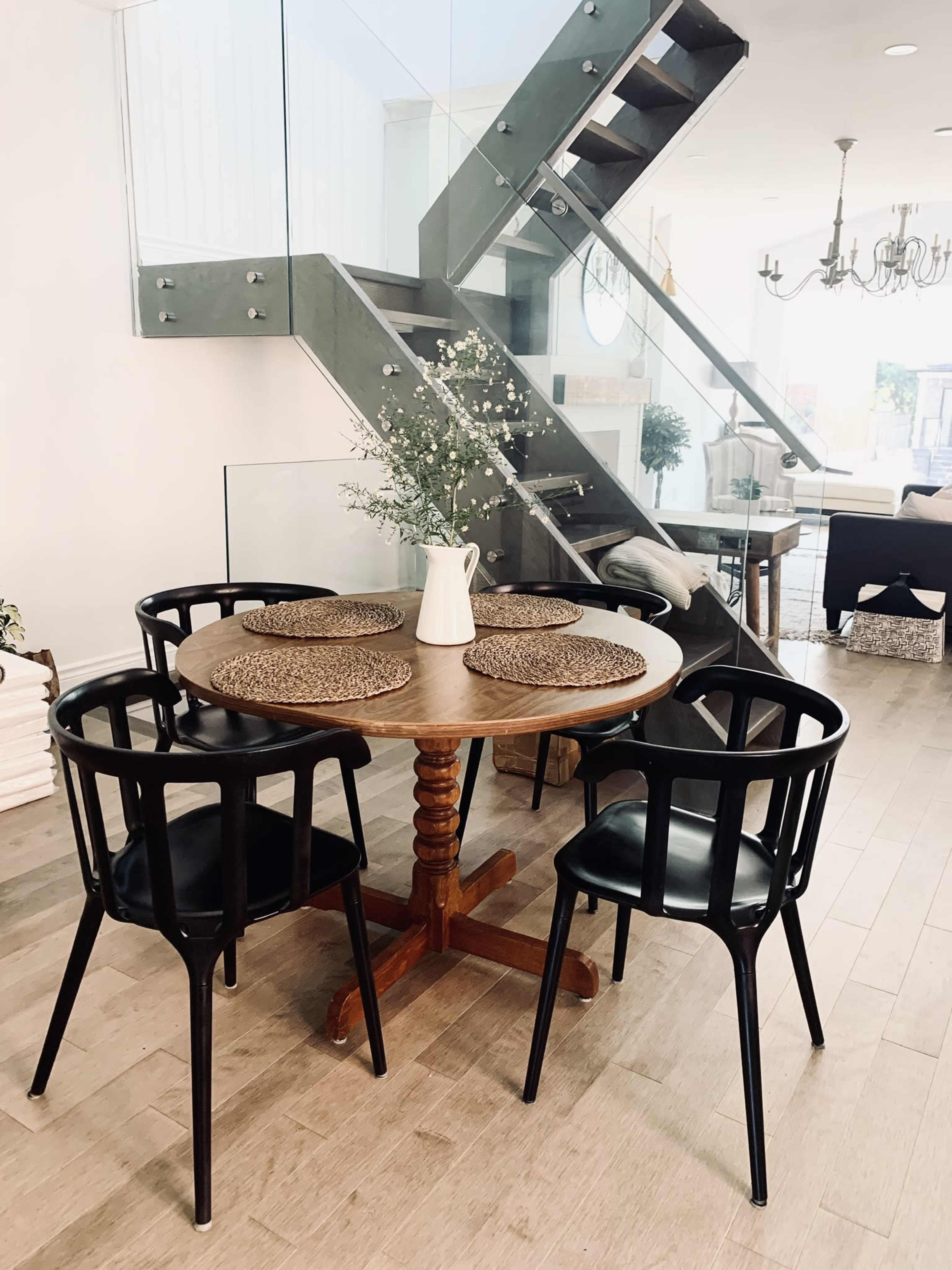 A round wooden dining table with four black chairs is arranged beneath a staircase with glass railing in a modern interior space.