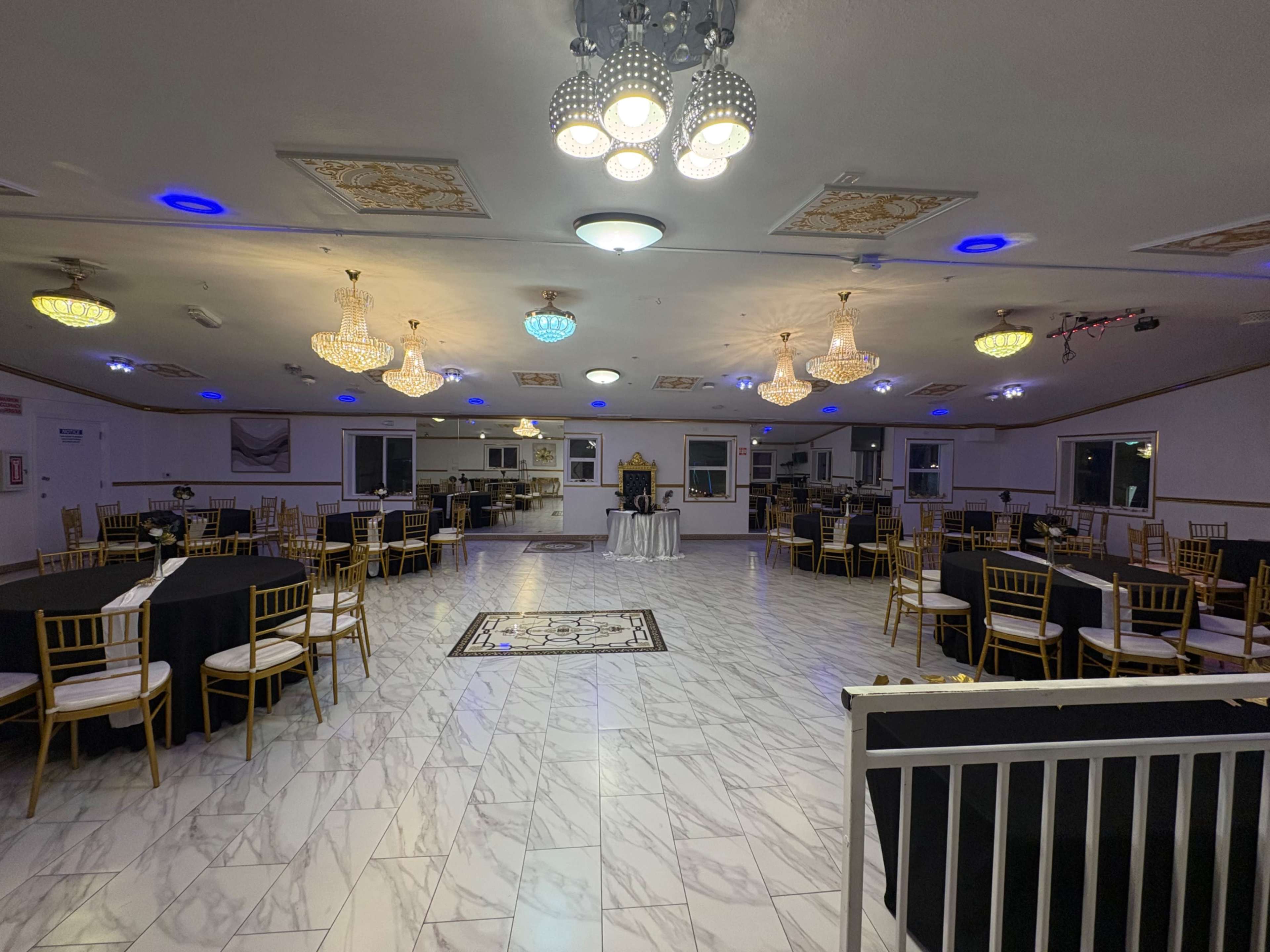 The image shows a spacious event hall with several round tables covered in black tablecloths, adorned with golden chairs and illuminated by various decorative ceiling lights.