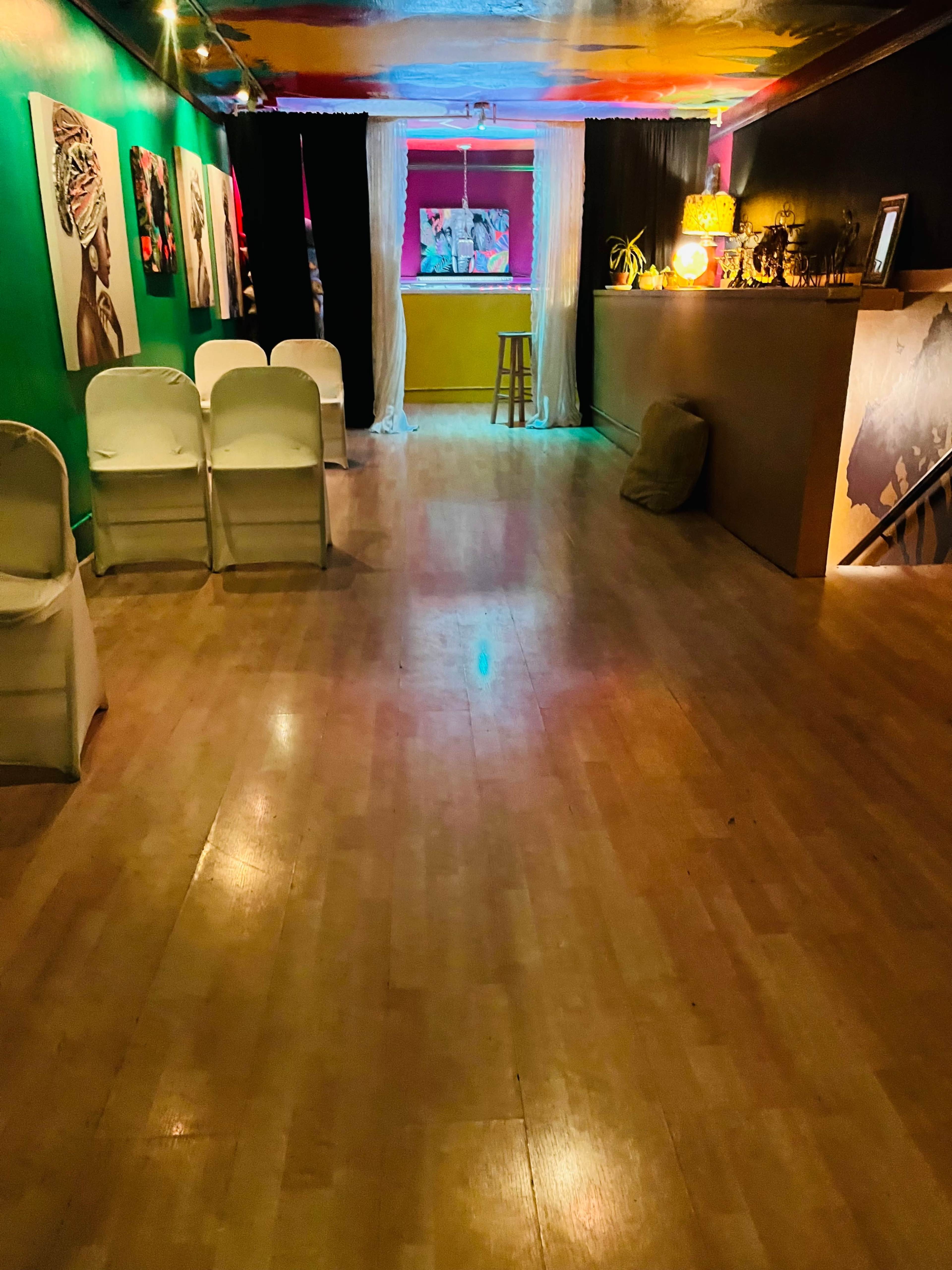 A sparse room with colorful walls, a small bar area, several white chairs arranged in a row, and artwork displayed on the walls.