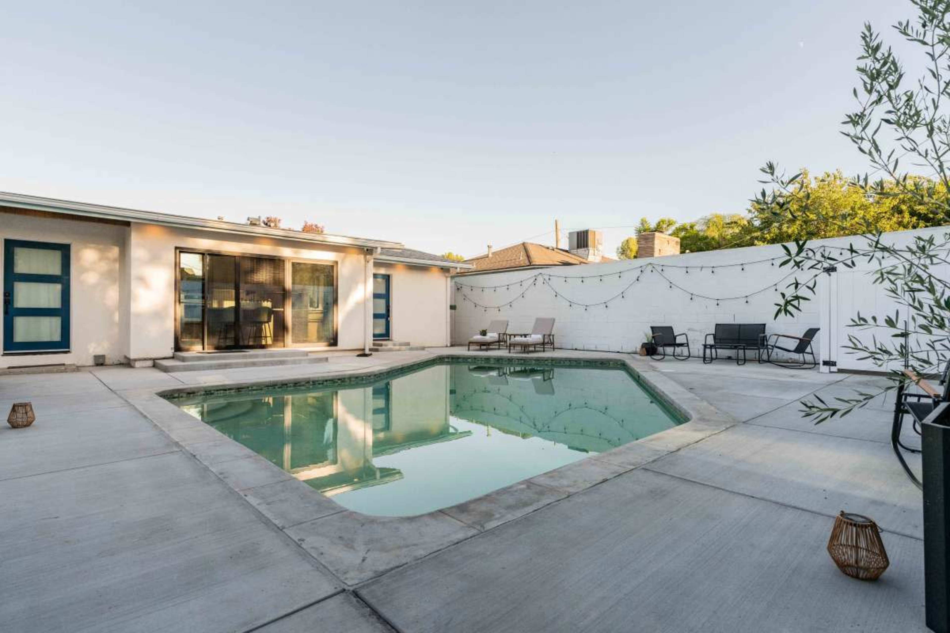 The image shows a backyard with a swimming pool surrounded by concrete, lounge chairs, and string lights hanging around a white wall.