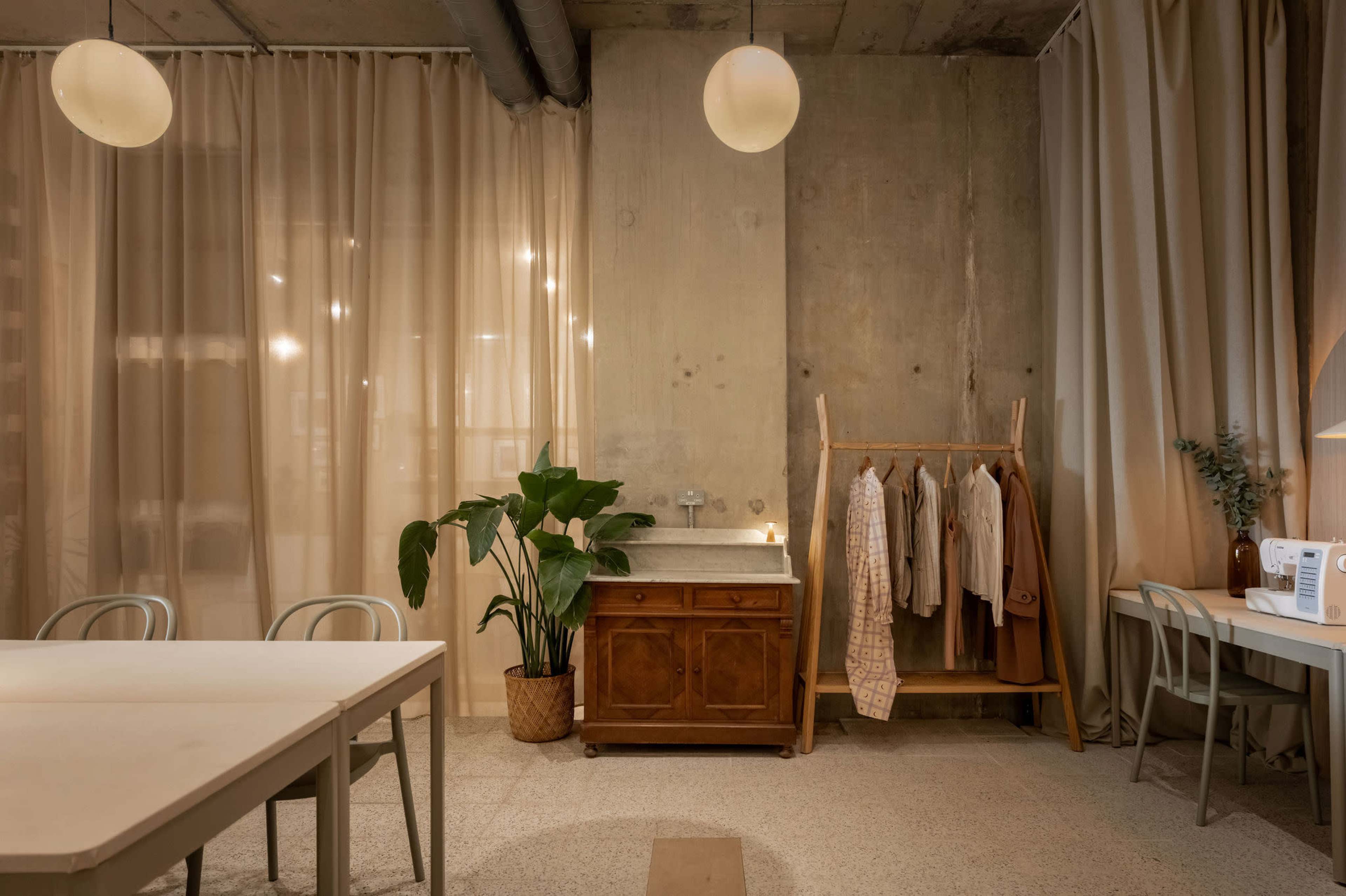 The image shows a modern interior space featuring a wooden dresser, a clothing rack with garments, a plant, and a dining table, surrounded by soft drapes and warm lighting.