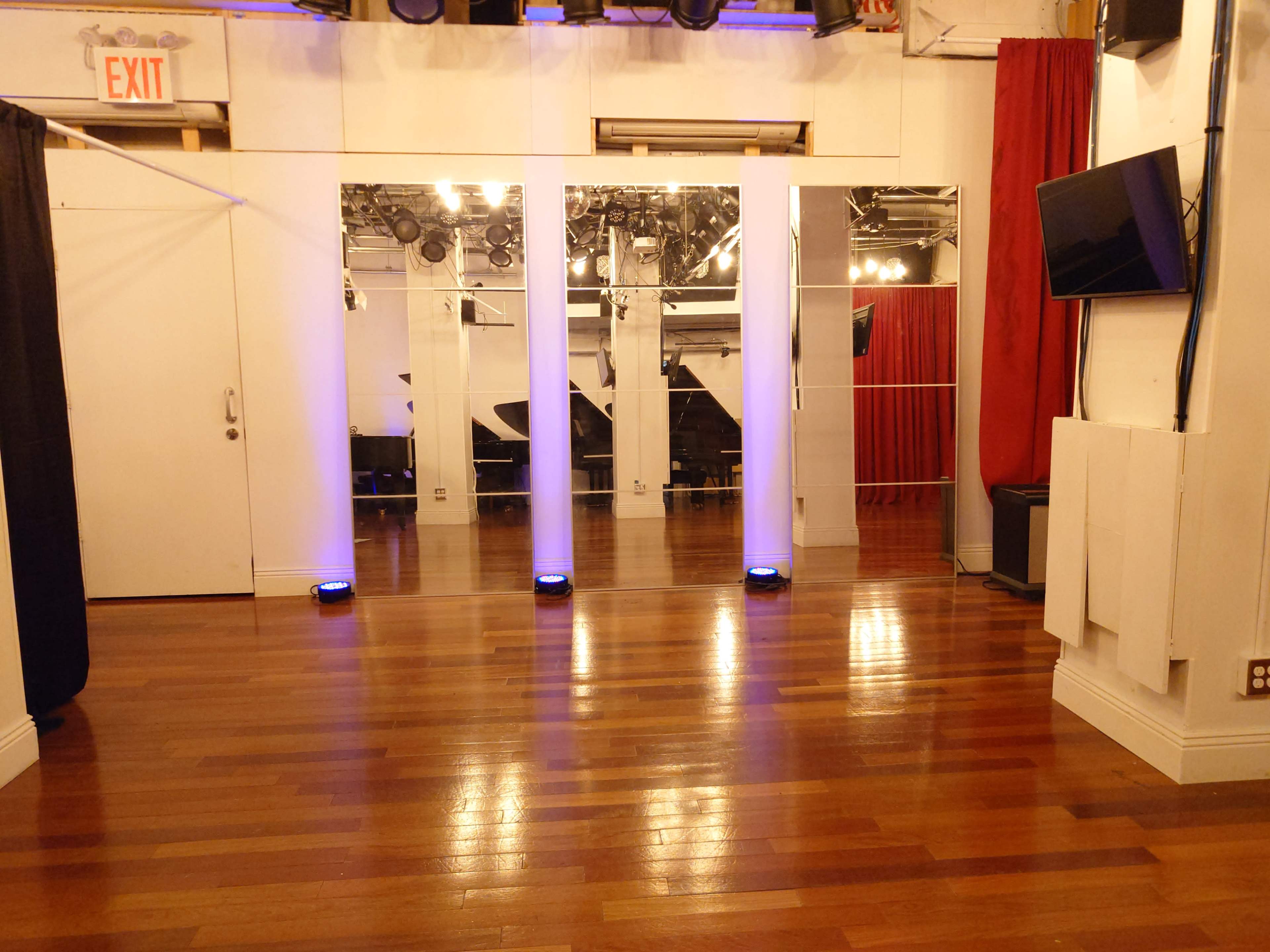 The image shows a spacious room with polished wooden floors, featuring tall mirrors, lighting fixtures, and a large black piano in the background.