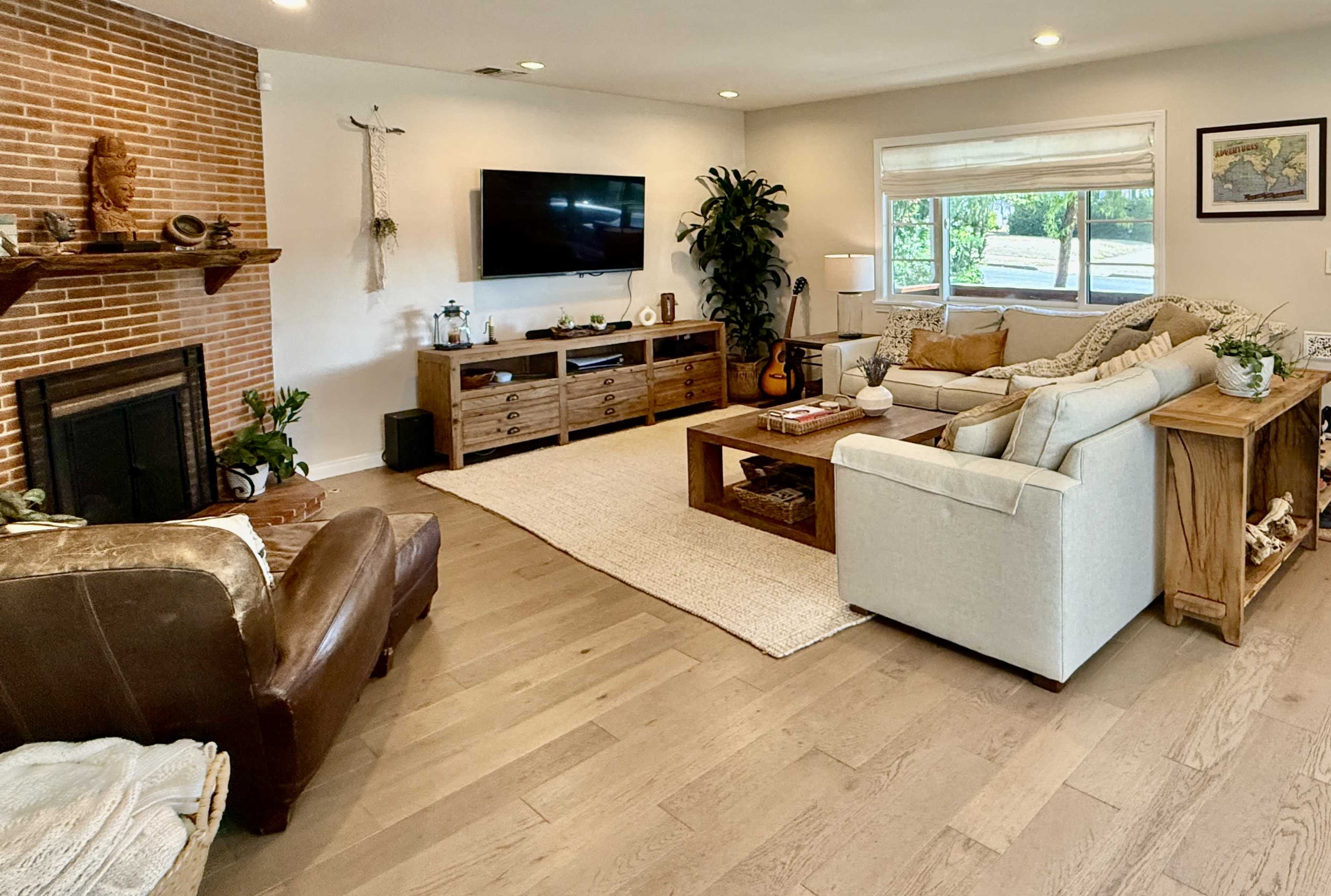 The living room features a cozy arrangement with a white sofa, a brown armchair, a wooden coffee table, and a large television mounted on the wall, surrounded by natural light from the windows.