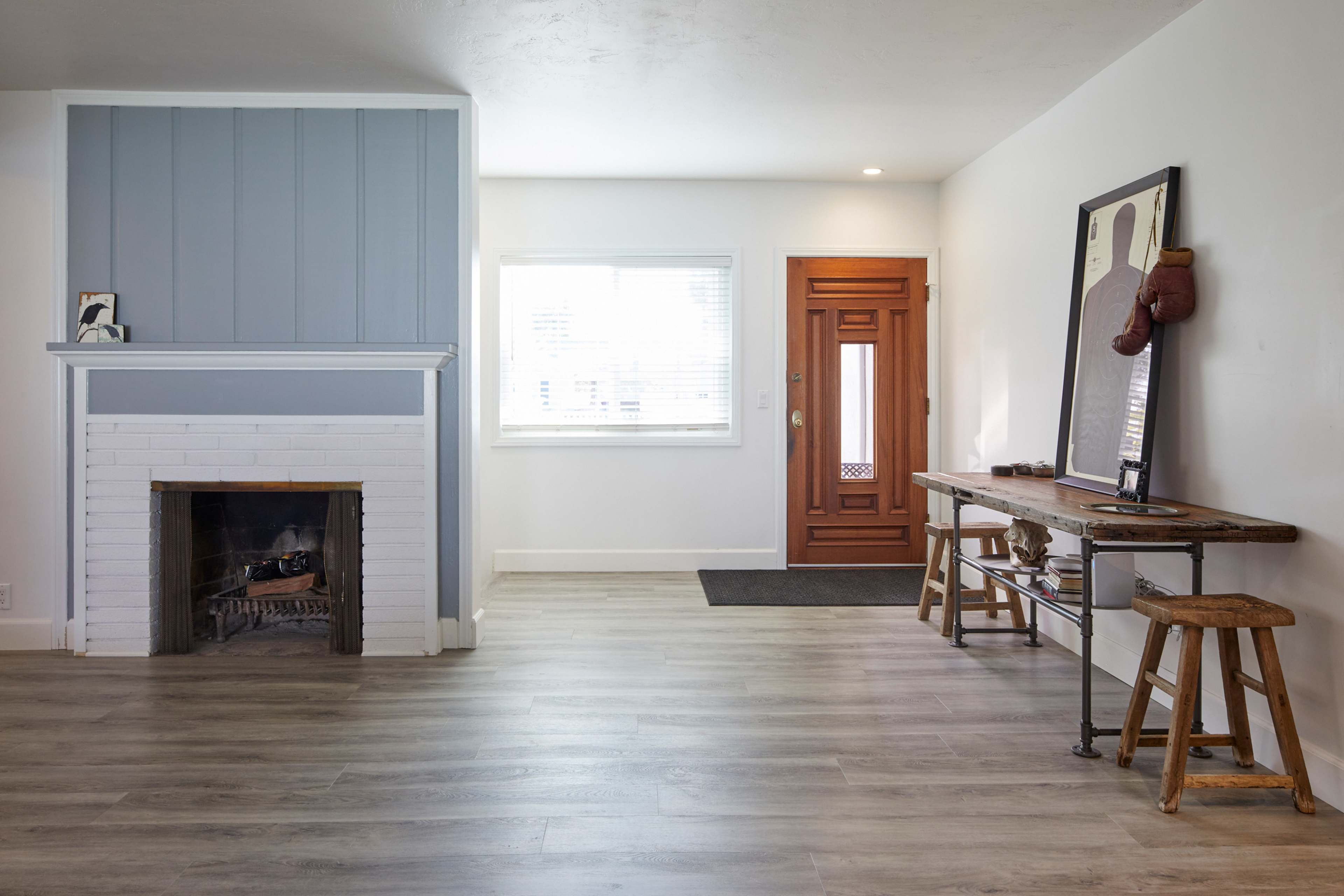 The image shows a minimalist living room featuring a white brick fireplace, a wooden entry door, and a simple wooden table with stools.