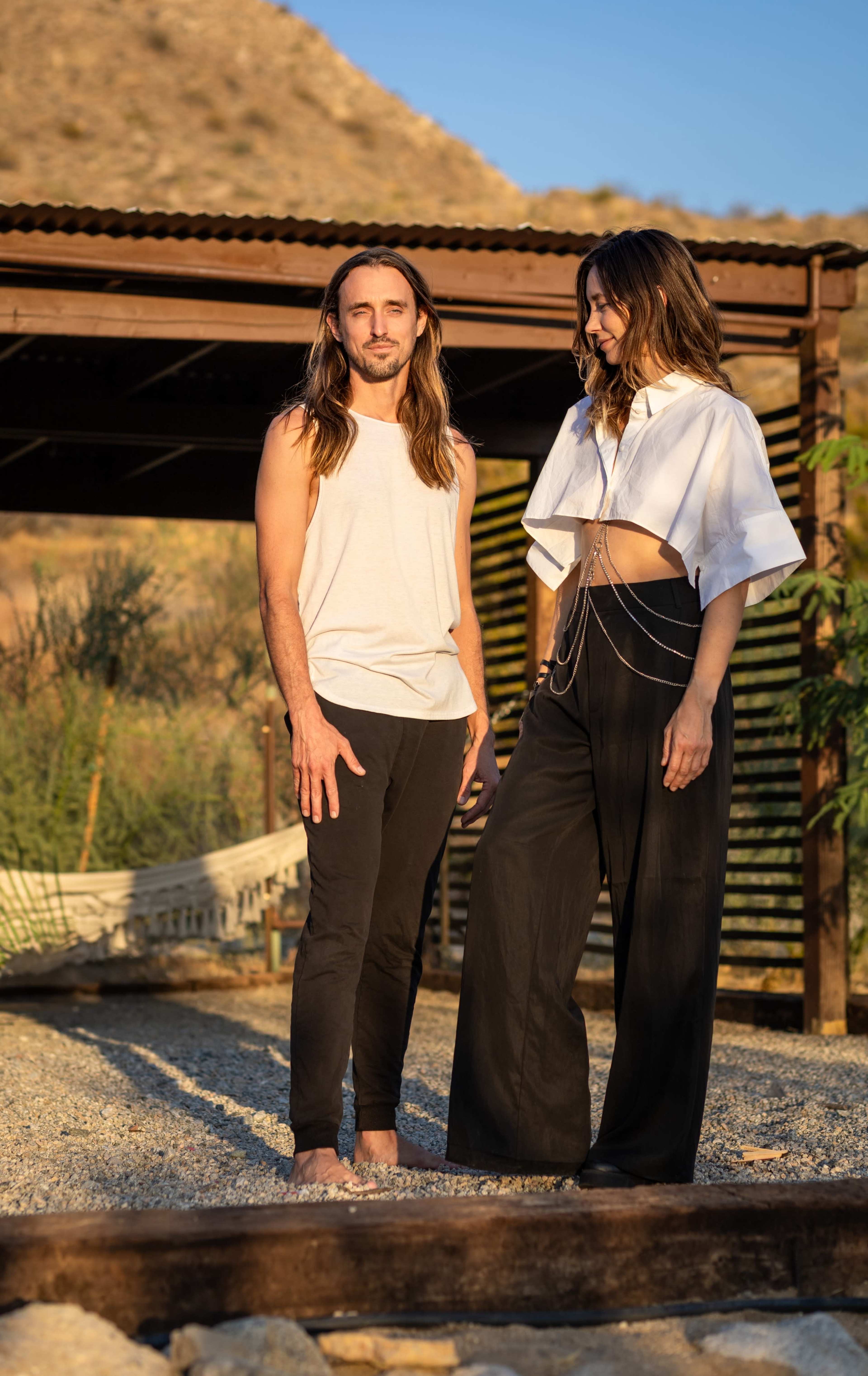 A man and a woman stand together outside near a wooden structure, surrounded by greenery and gravel.