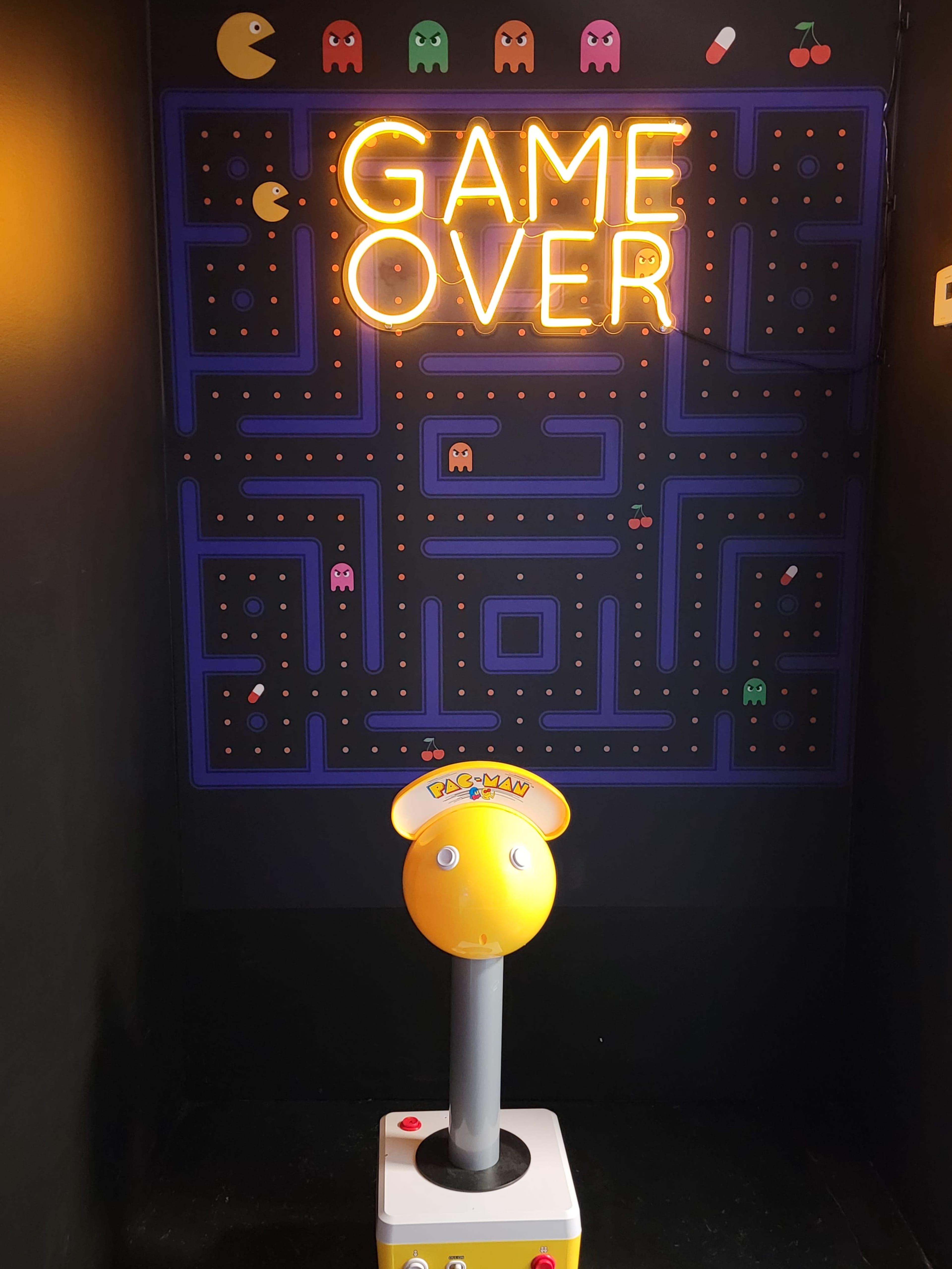 The image features a neon sign reading "GAME OVER" against a black wall adorned with a Pac-Man maze and a cartoon character head on a pedestal.