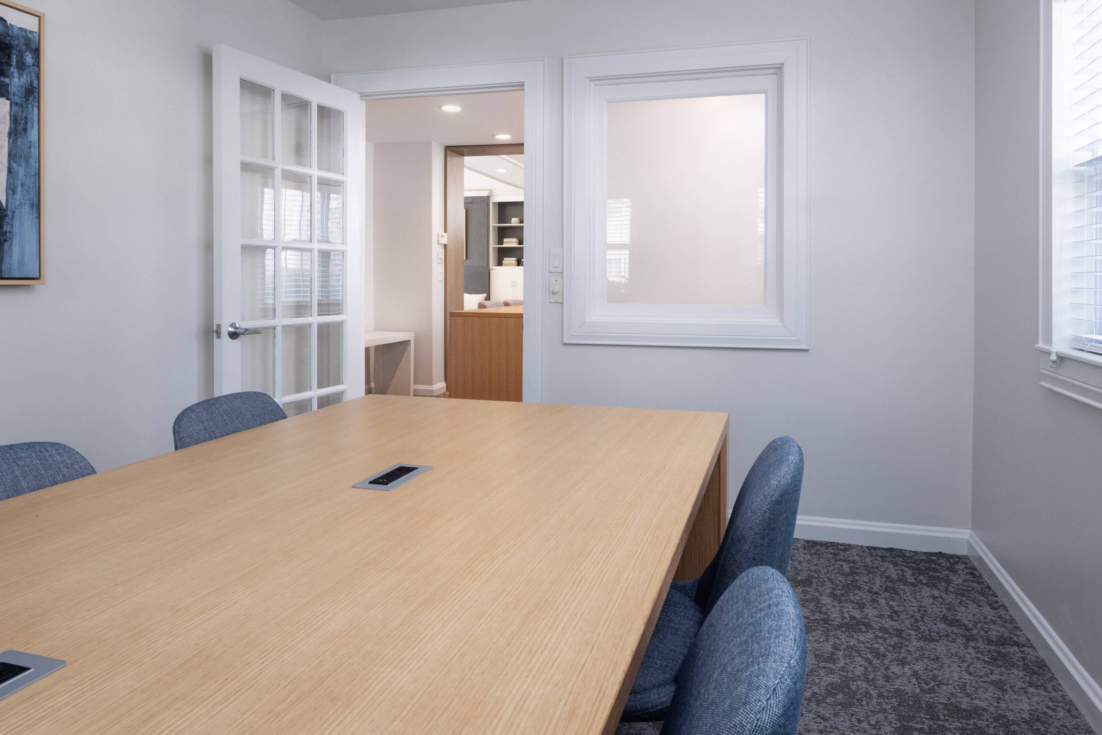 A conference room features a large wooden table surrounded by blue chairs, with a doorway leading to a nearby office space.